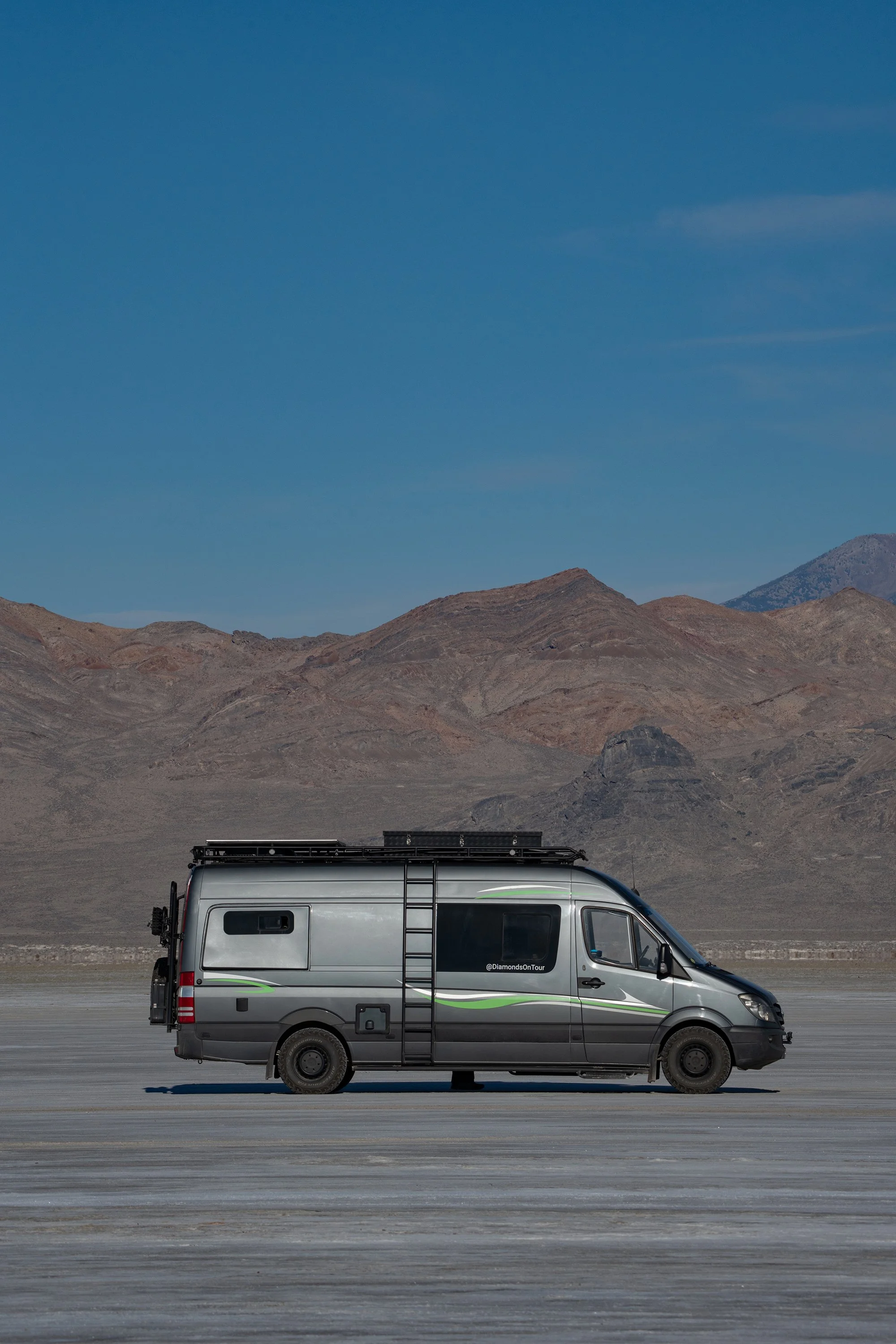 A silver van with green and black decals driven on a flat, dry landscape with mountains in the background and a mostly clear blue sky. Bonneville Salt Flats.