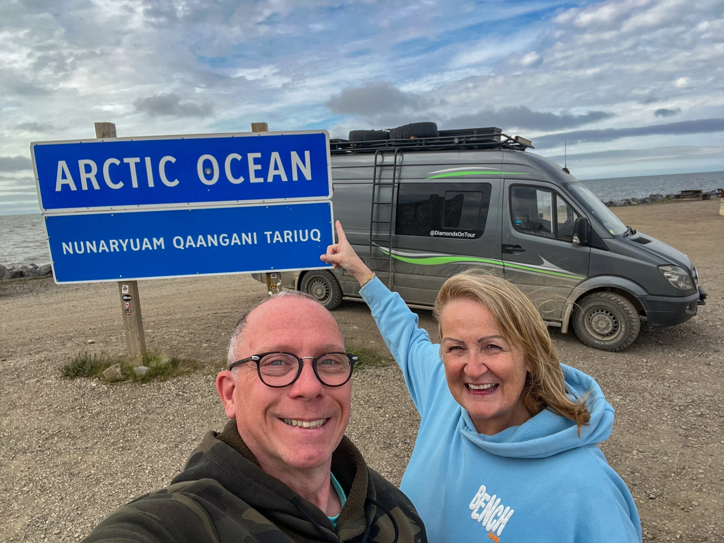 Two people smiling for a selfie at the Arctic Ocean, with a sign indicating the location and a van parked on a dirt area near the shore, with cloudy sky and ocean in the background.