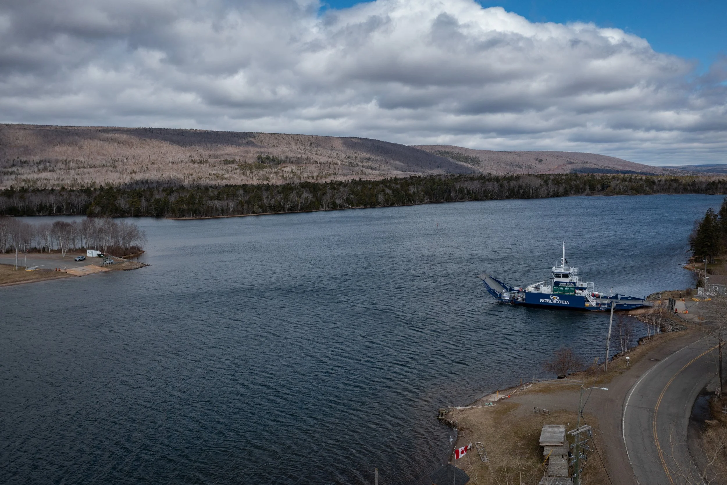 A large harbor with a ferryboat named Nova Scotia docked at the shore. The water is surrounded by forested hills and cloudy sky.