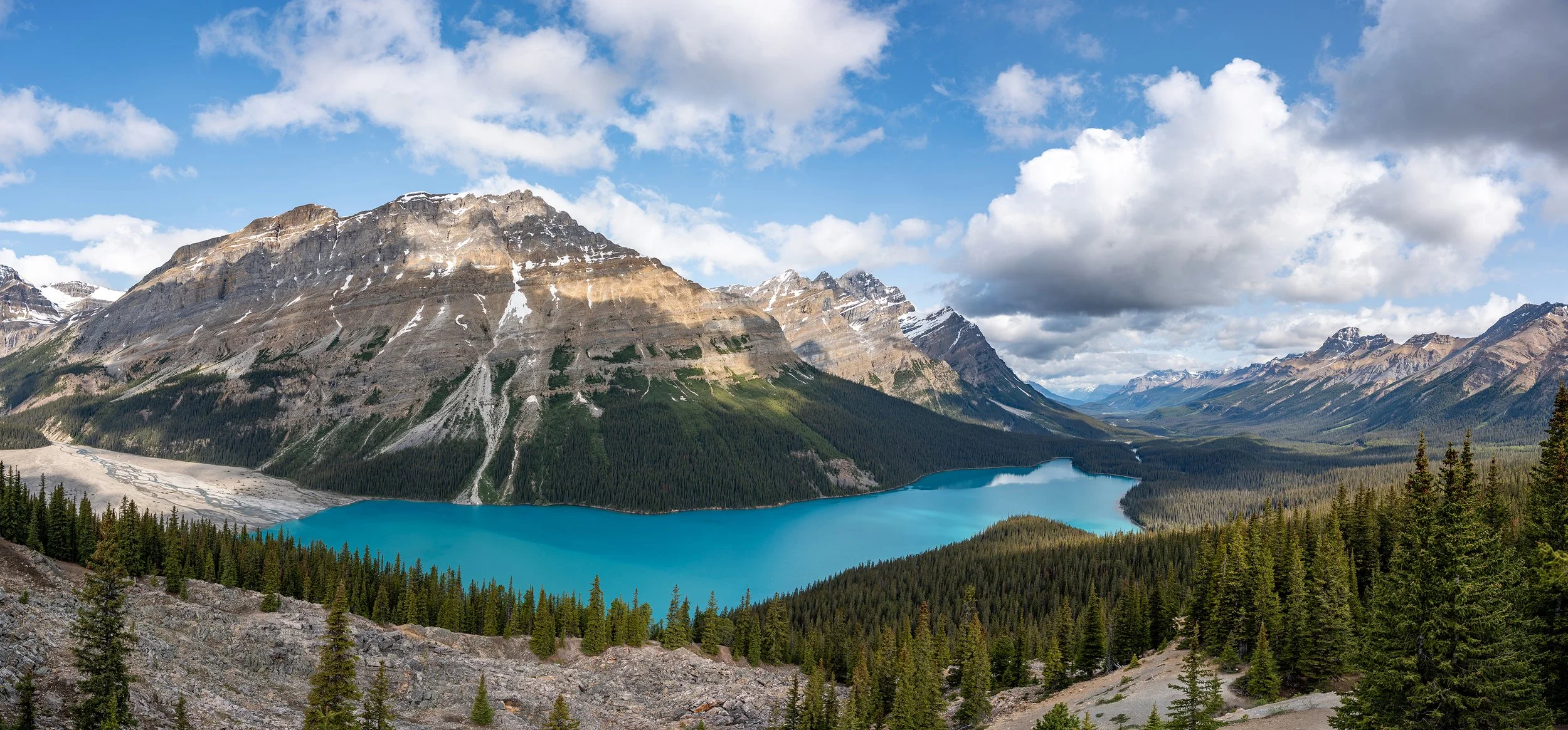 A scenic view of a turquoise mountain lake surrounded by green pine forests and rugged mountains with patches of snow.