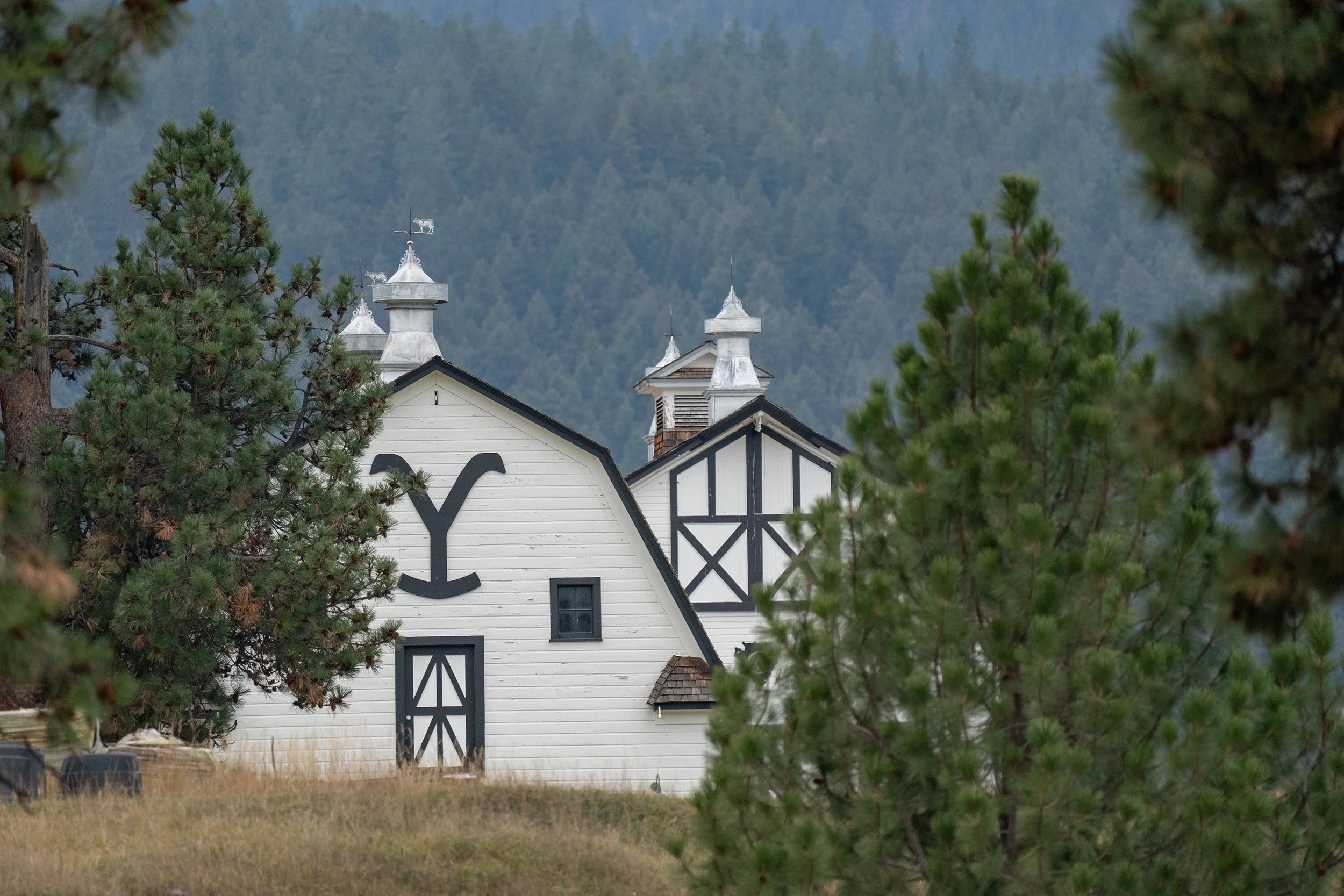 A white building with a black and white barn-style design, surrounded by pine trees with a mountain in the background. Yellowstone Dutton Ranch.
