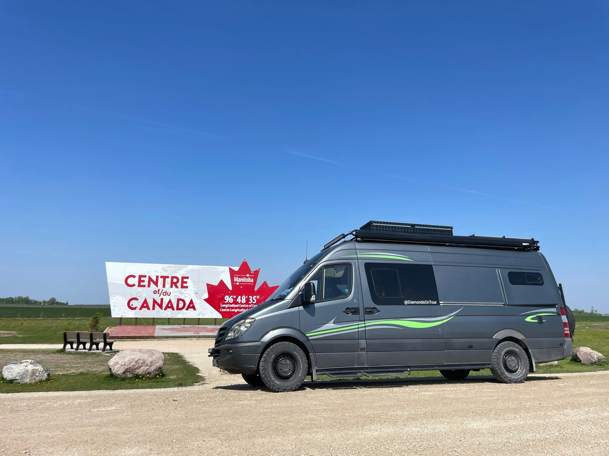 A grey van with green and black stripes parked on a grassy area near a walkway, with a sign that says 'Centre of Canada' and a red maple leaf logo behind it, under a clear blue sky.