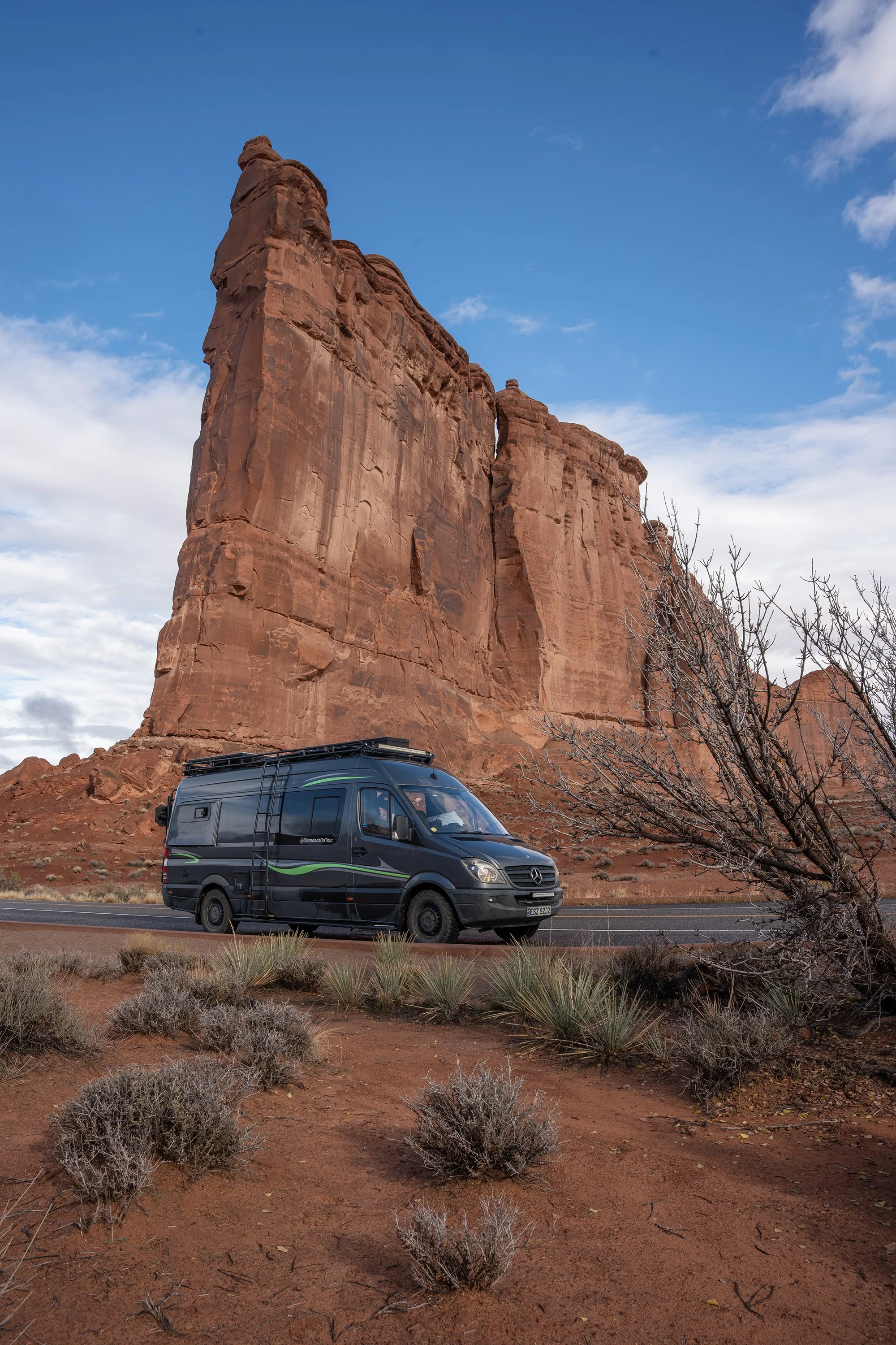 A black camper van parked on the side of a road in a desert landscape with large red rock formations and sparse bushes, under a partly cloudy sky.