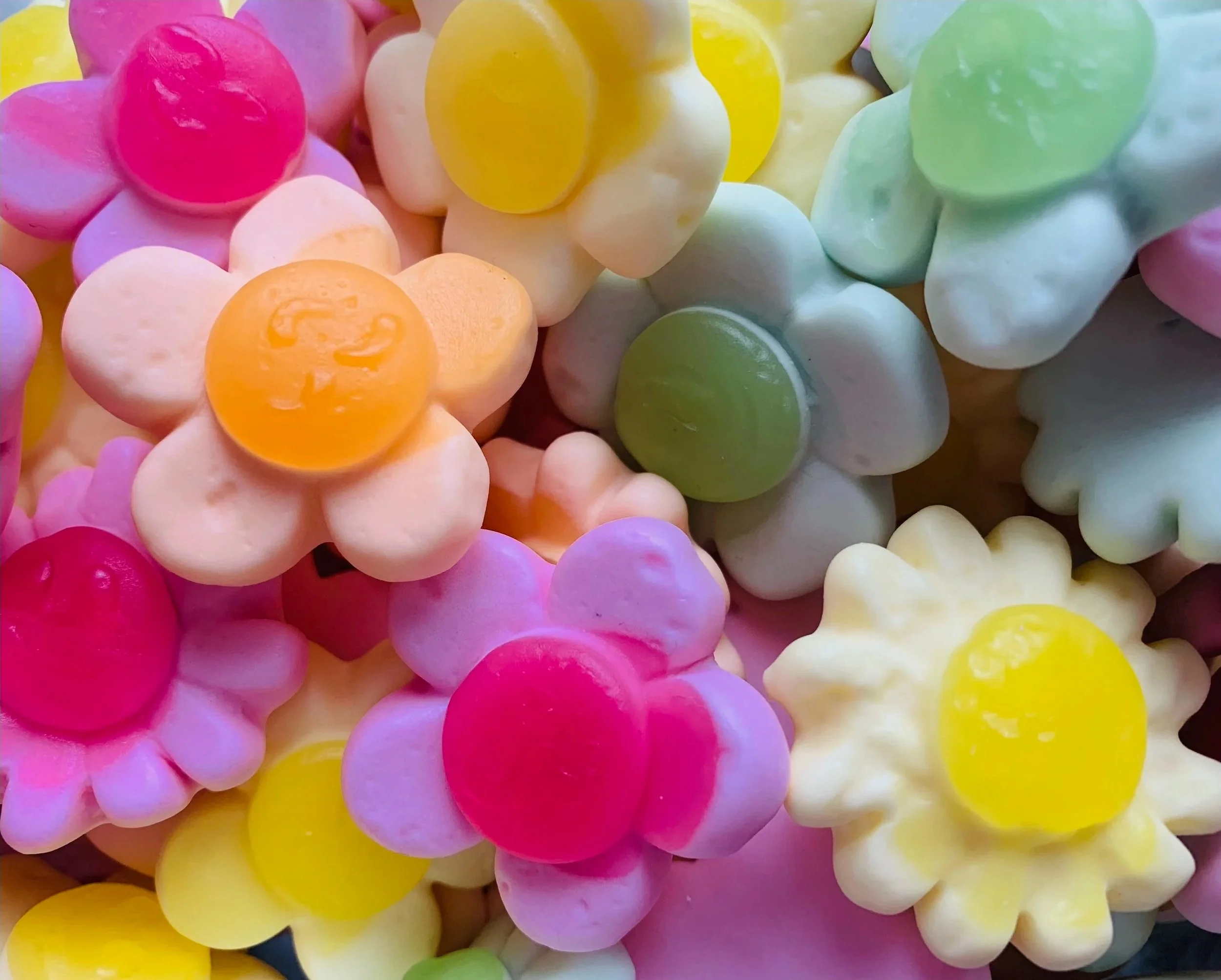A collection of colorful jelly candies in various pastel and bright colors, shaped like flowers with round centers, piled together.