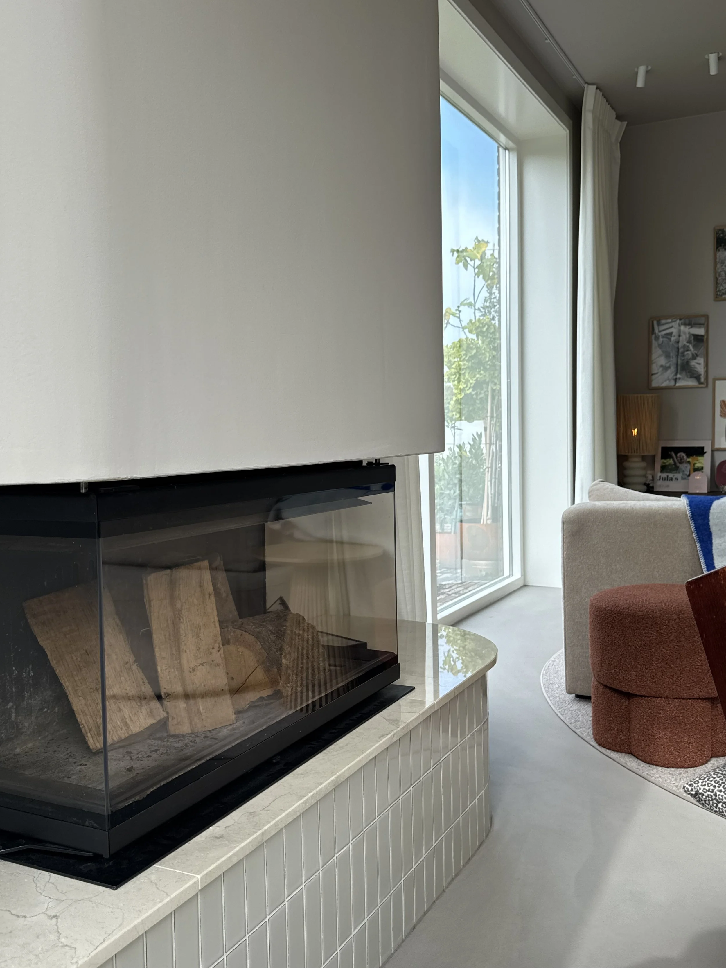 Living room with a window, beige sofa, red ottoman, and a fireplace with wood inside.
