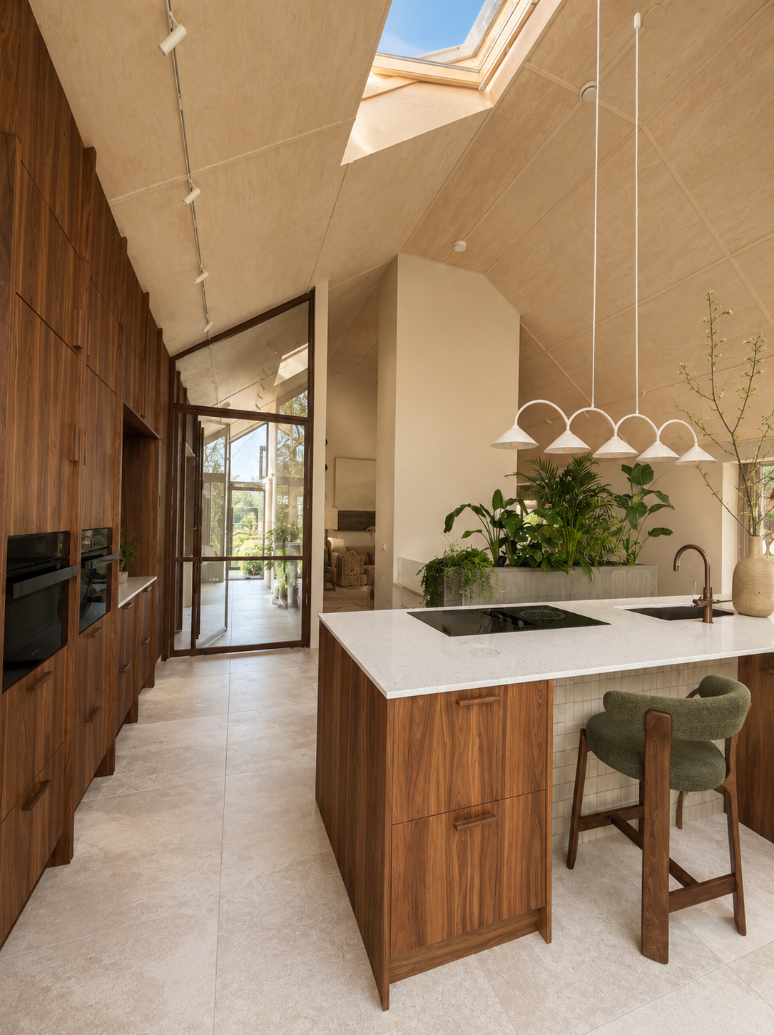 Modern kitchen with a central island featuring a white countertop, surrounded by wooden bar stools, green plants behind the island, and a dining area with white chairs near large windows with white curtains.