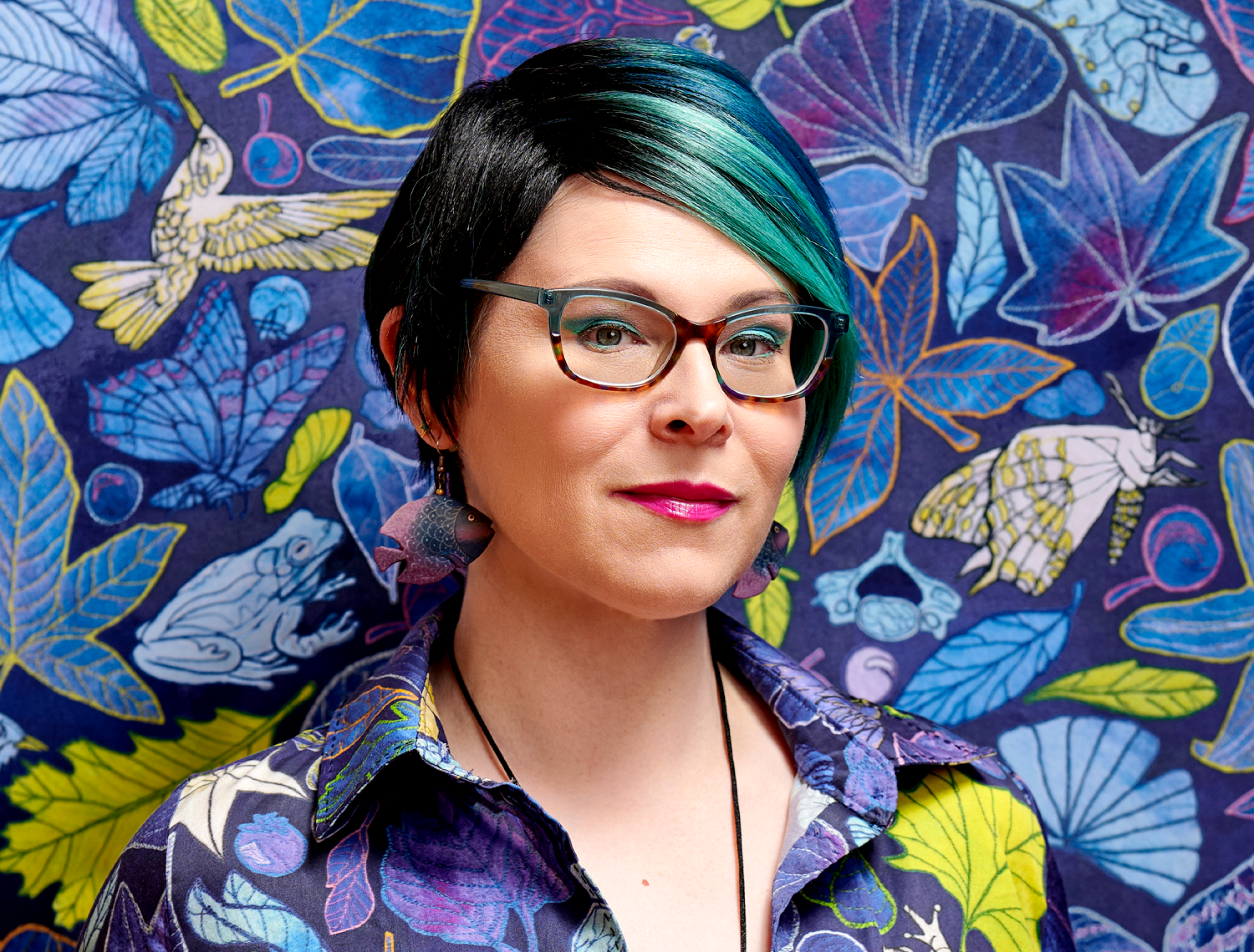 woman with teal hair and glasses stands in front of wallpaper which matches her shirt