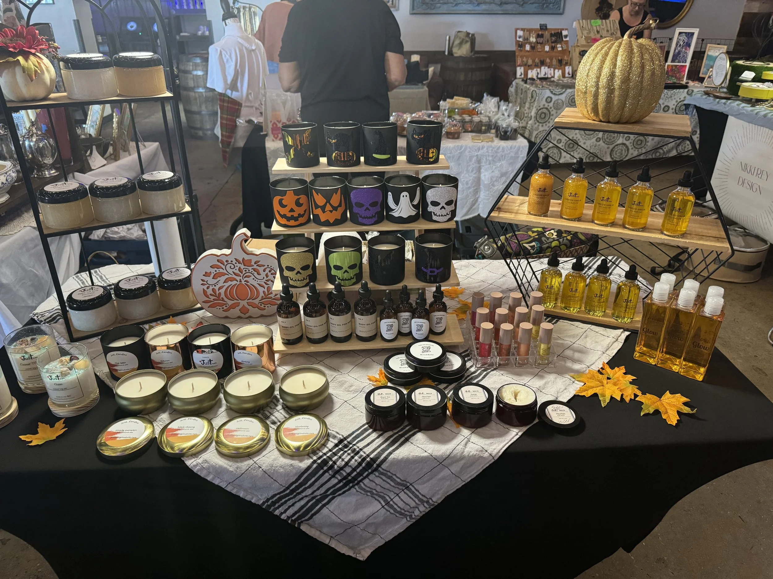 Table display of Halloween-themed candles, mugs with pumpkin, skull, ghost, and jack-o'-lantern designs, various bottles of scented oils, lip glosses, and decorative autumn leaves at a craft market.