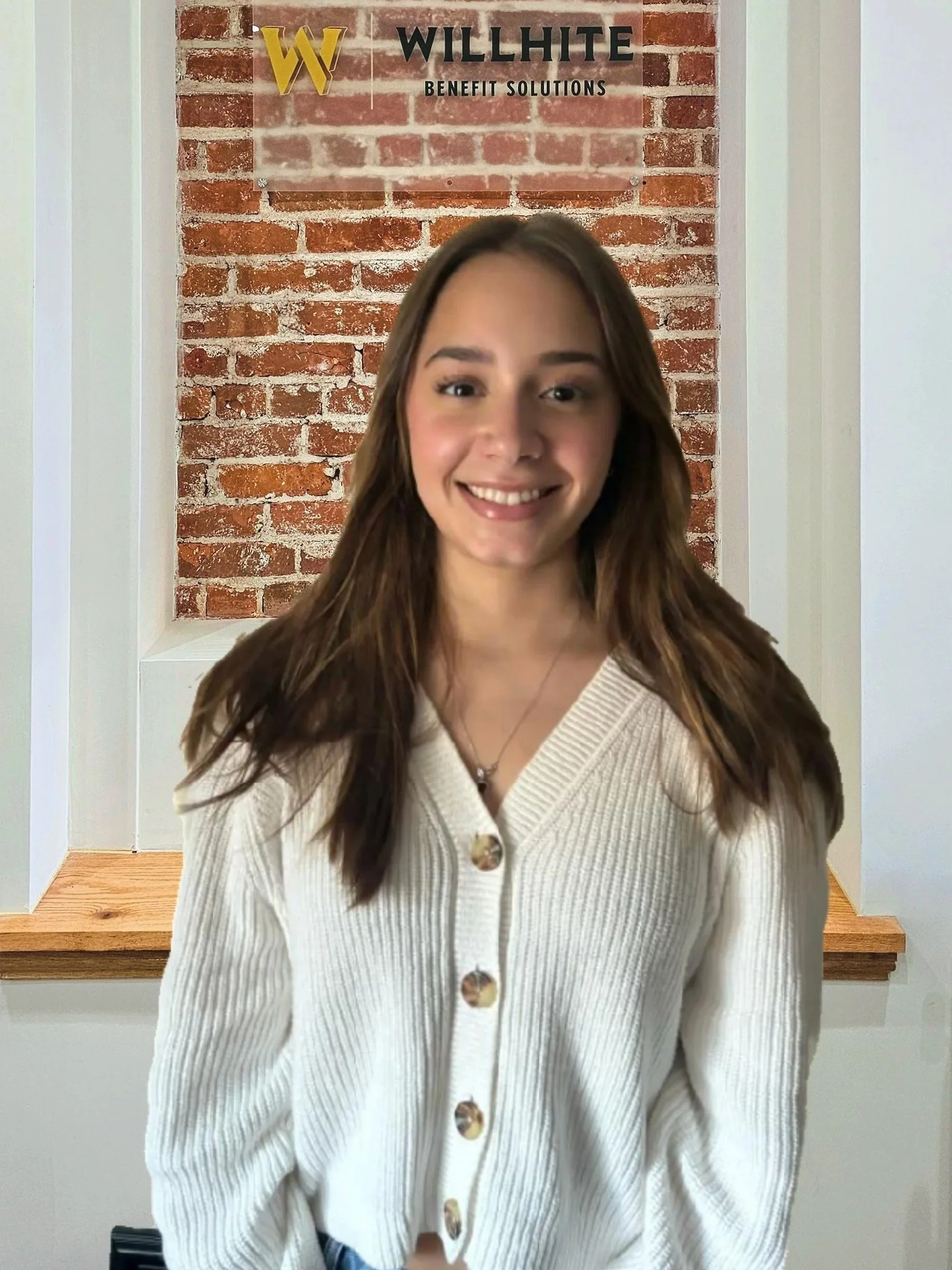 A young woman with long brown hair, smiling, wearing a white button-up cardigan, standing indoors in front of a brick wall with a sign that reads 'WILLHITE BENEFIT SOLUTIONS'.