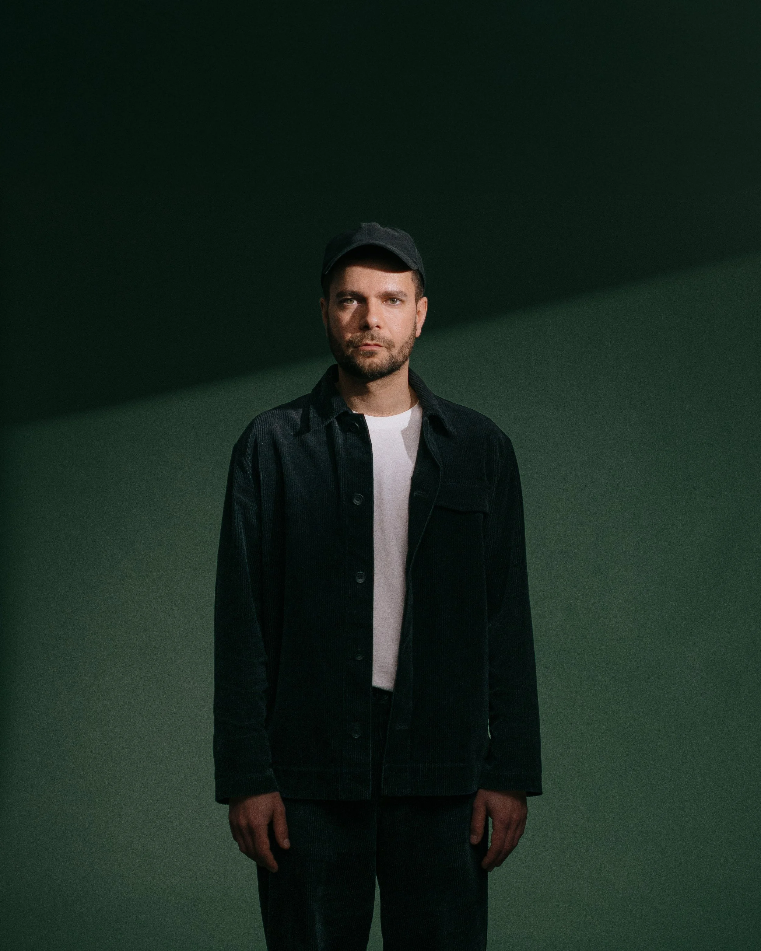 Junge Mann trägt schwarze Jacke, weißes T-Shirt und schwarze Mütze, steht vor dunklem grünen Hintergrund.