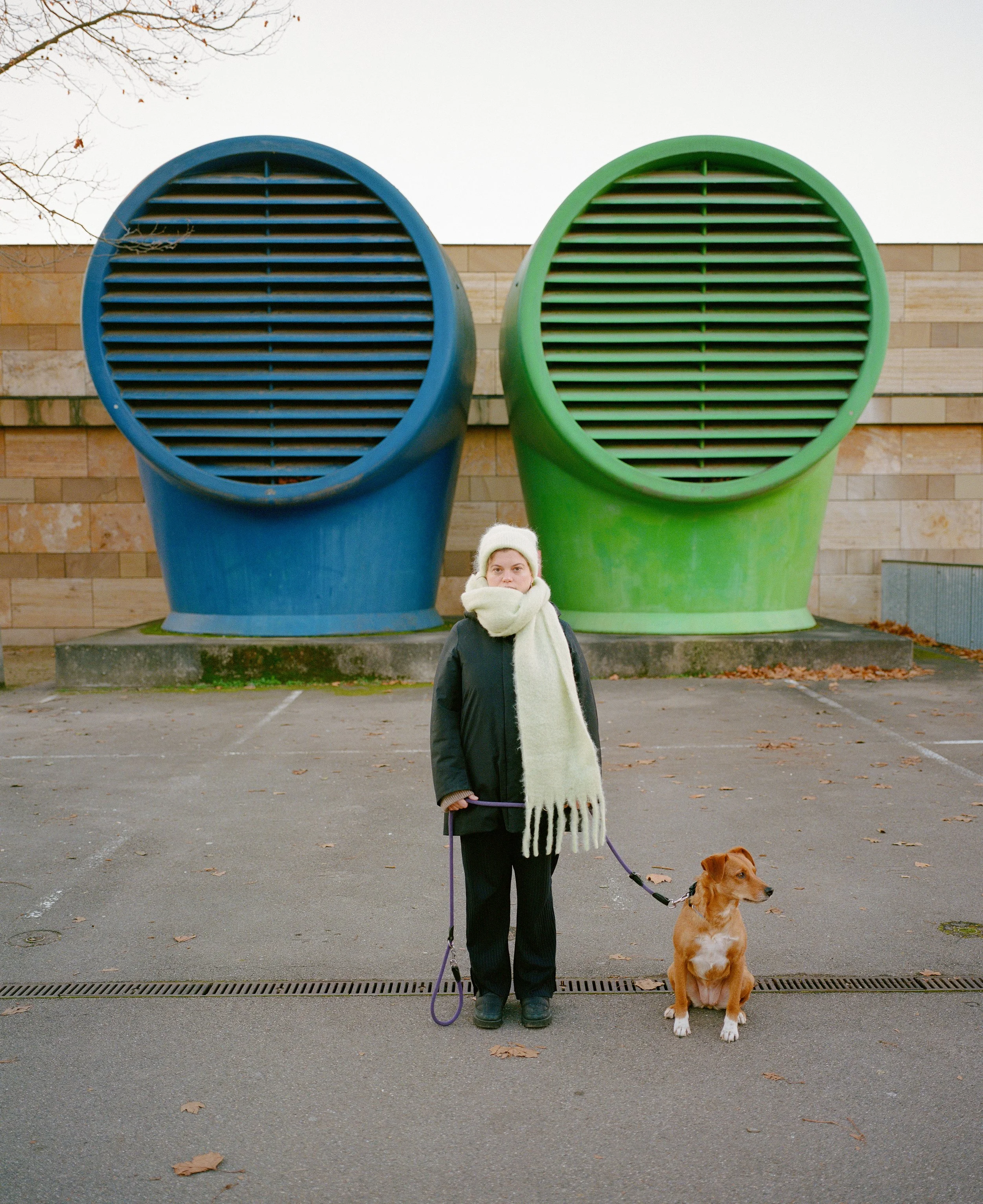 Person mit Schal und Mütze hält einen Hund an der Leine vor zwei großen farbigen Lüftungsrohren, blau und grün, auf einem Parkplatz.