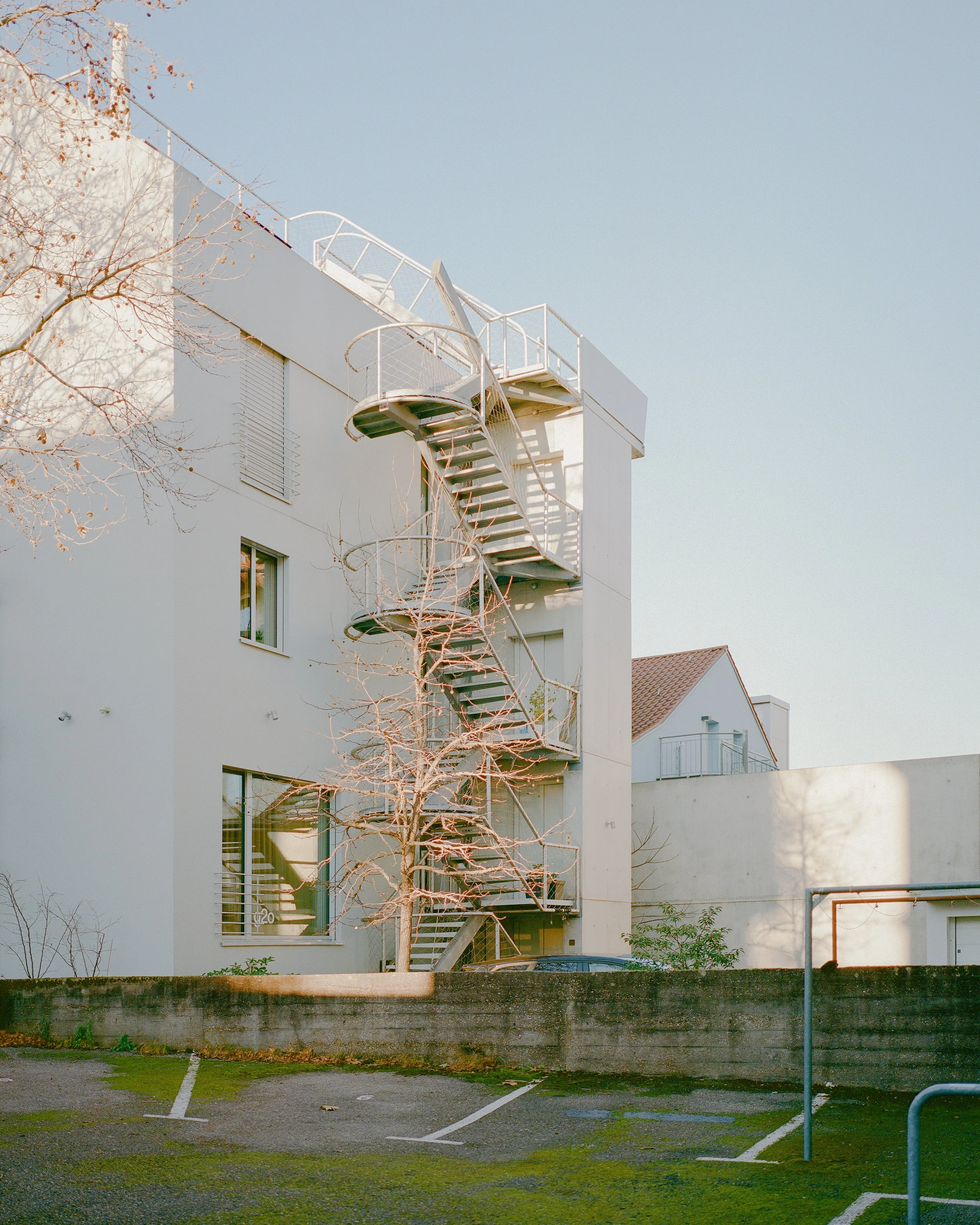 Ein modernes Gebäude mit einer Wendeltreppe an der Außenseite, ein kahler Baum im Vordergrund, daneben ein leerer Parkplatz.