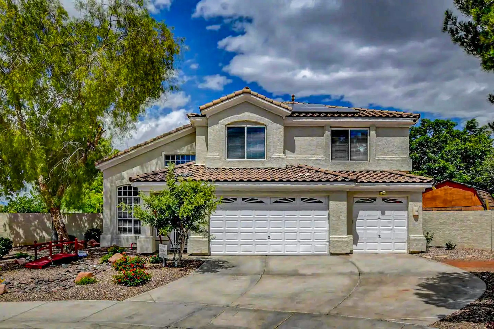 Two-story stucco house with tiled roof, three-car garage, and landscaped front yard featuring trees and shrubs, set against a blue sky with clouds.