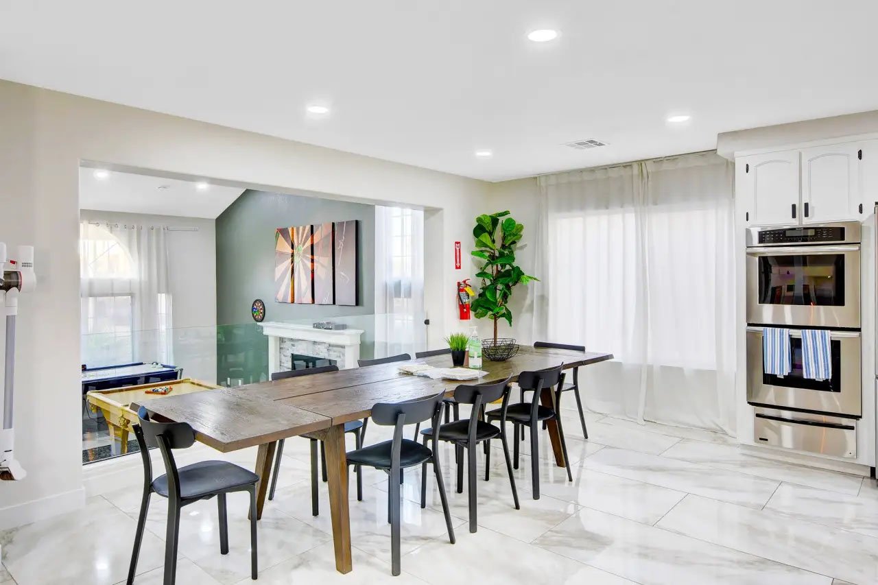 Modern kitchen and dining area with a wooden table, six chairs, double oven, green plant, and large windows with curtains.