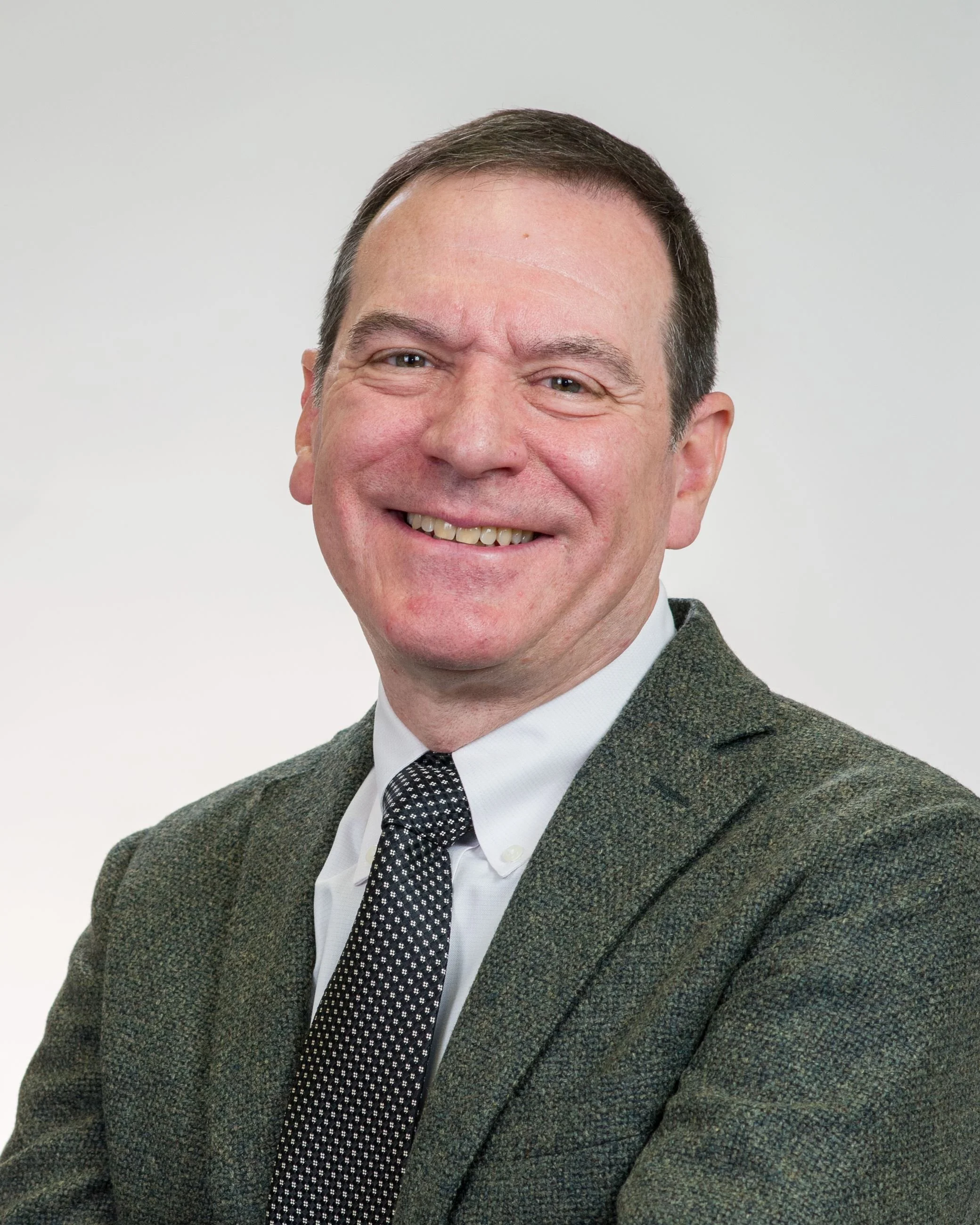 A smiling man wearing glasses and a suit with a white shirt, standing outdoors with a background of a brick building and windows.