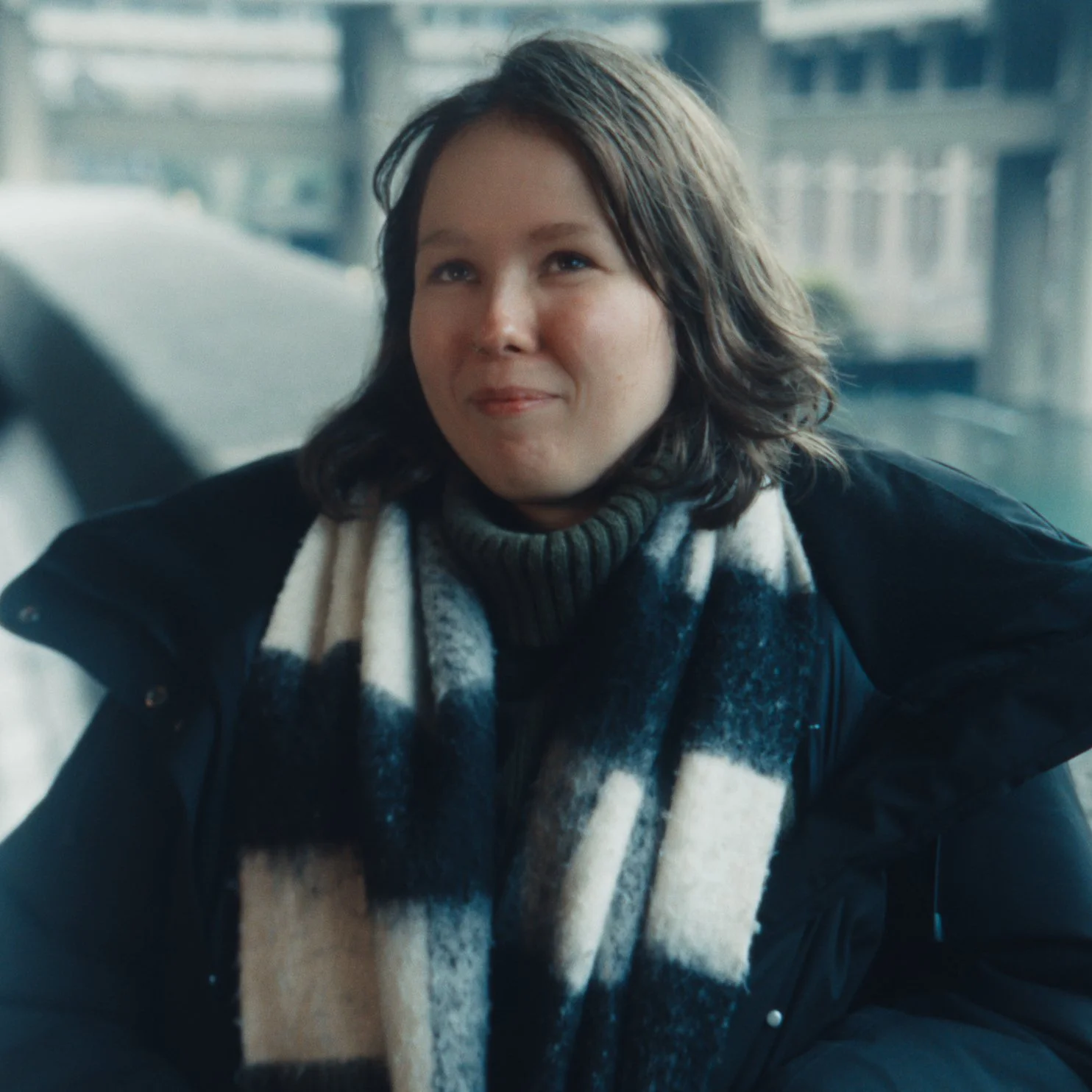 A woman with short brown hair, wearing a black coat and a black, white, and gray checked scarf, sitting on a bus or train with a window behind her showing a blurred cityscape.