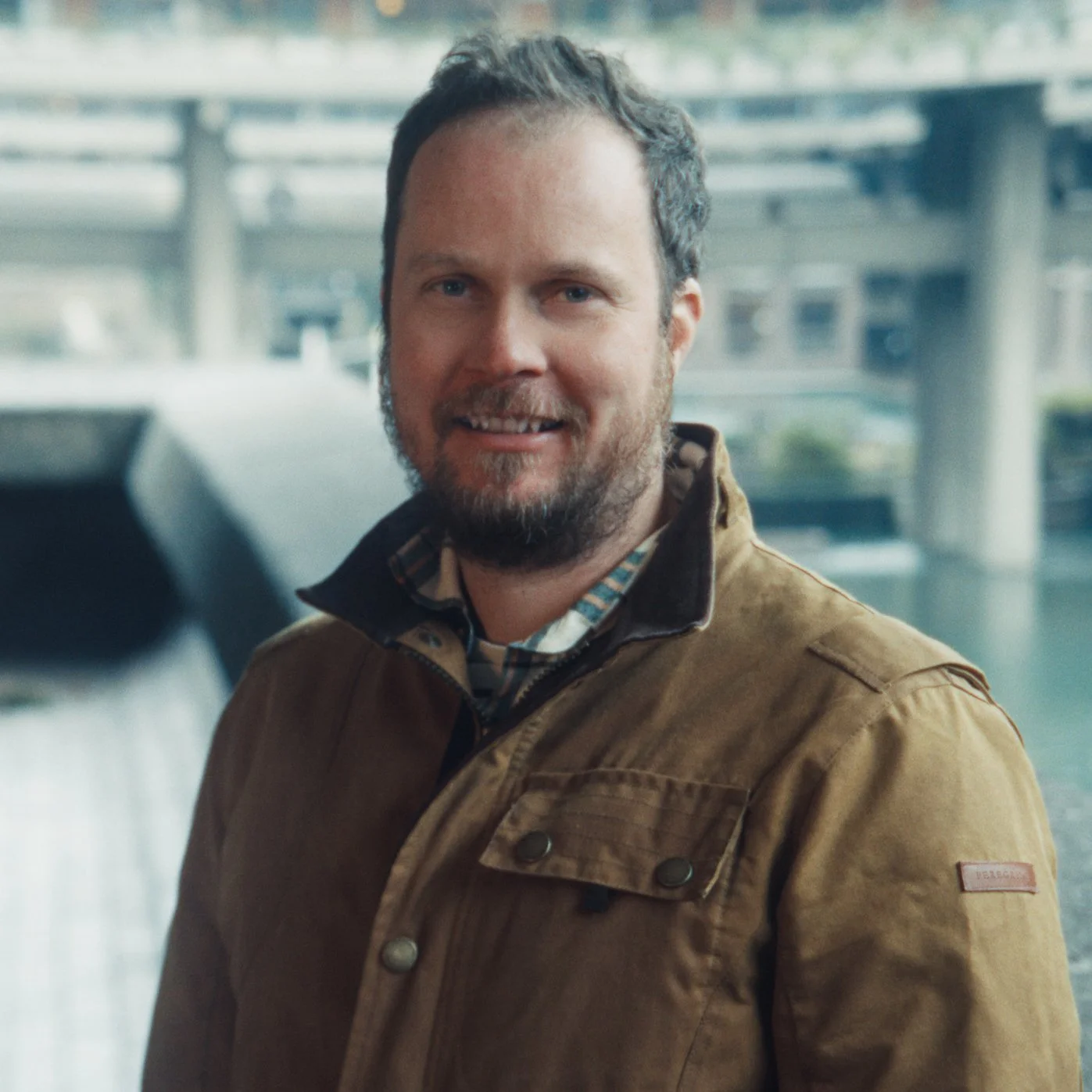A man with a beard smiling outdoors in front of a blurred background of a bridge and water.