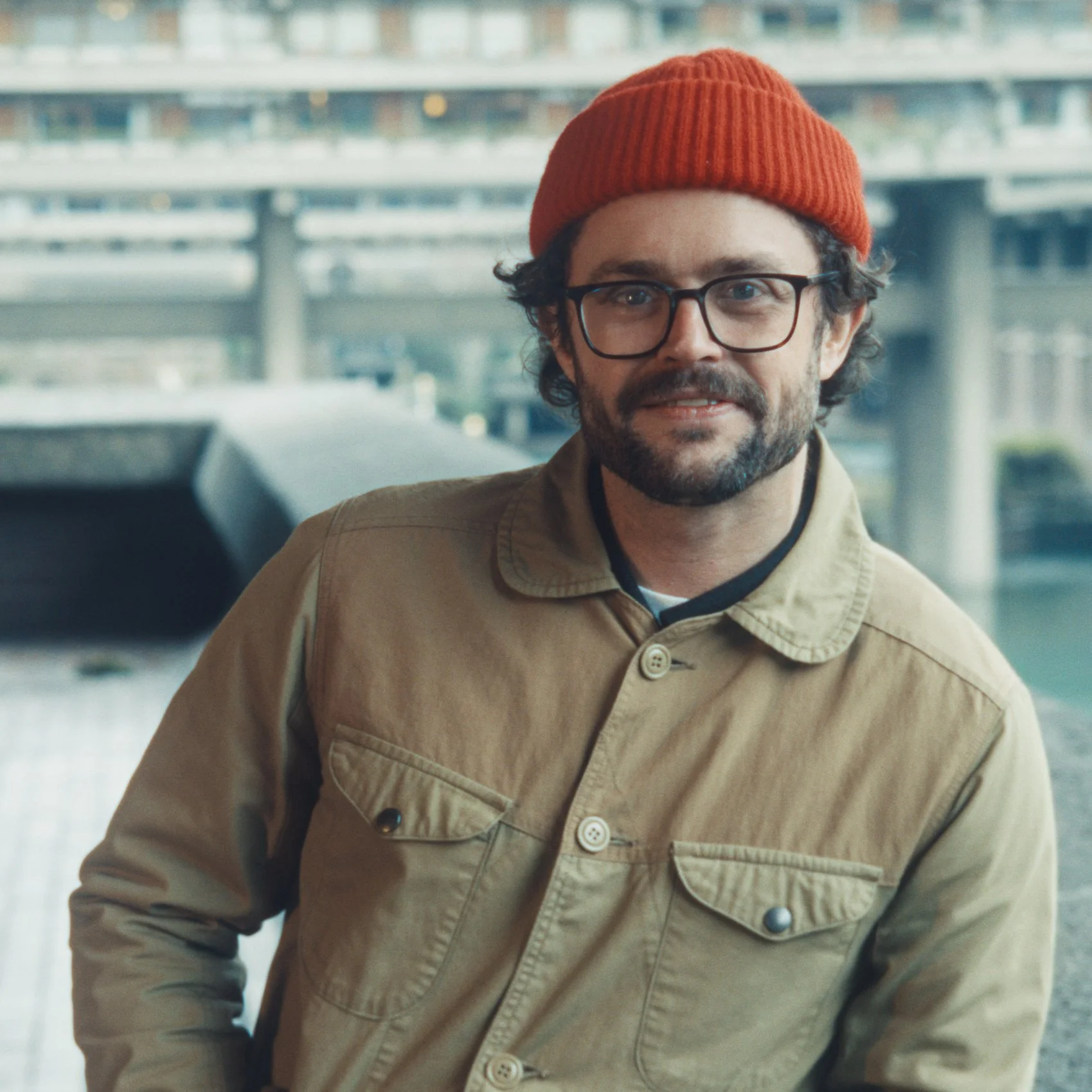 A man with glasses, dark hair, and a beard wearing a red knit beanie and a beige jacket, smiling and looking at the camera, indoors with a blurred background of a multi-story parking garage.