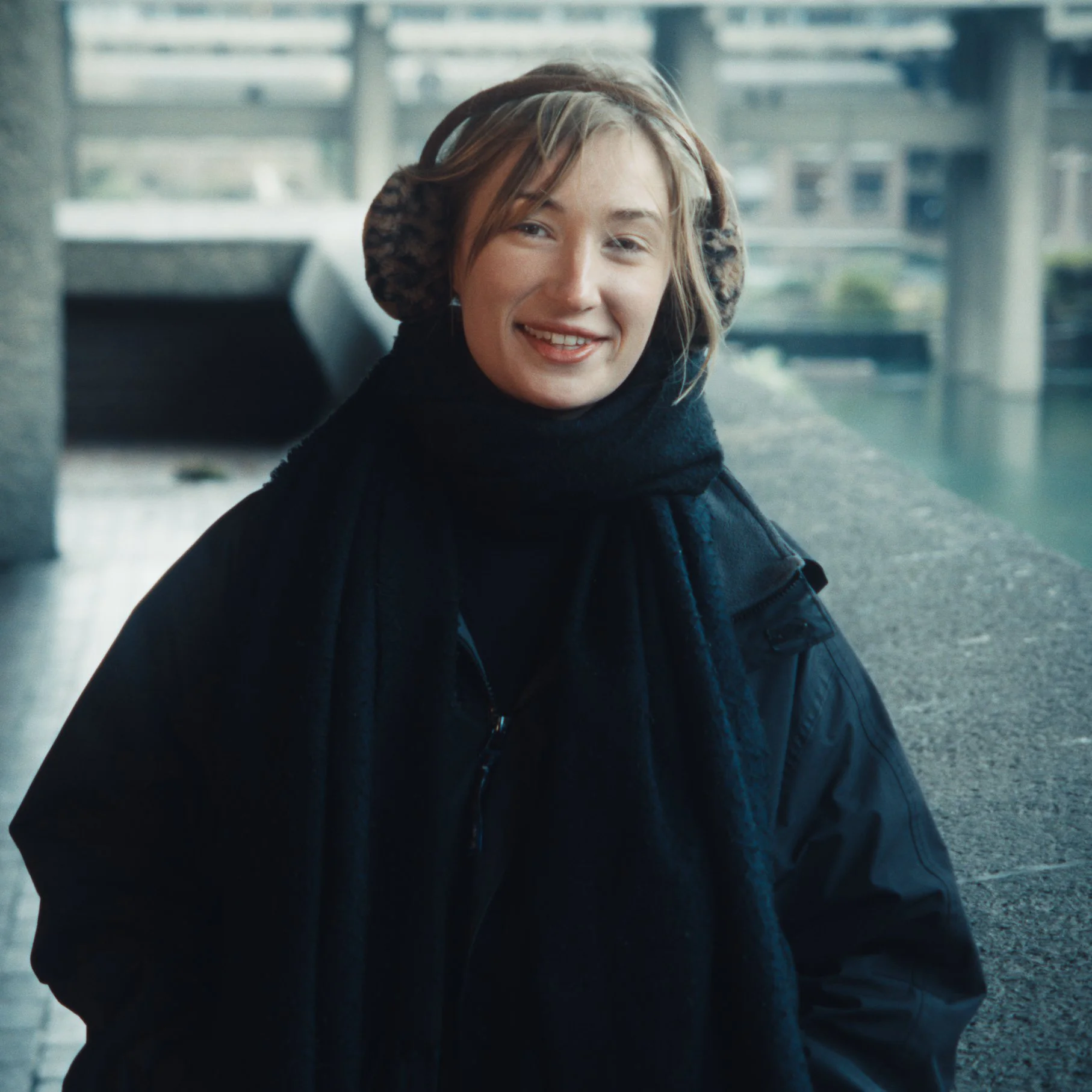 A young woman smiling, wearing headphones and a black jacket, standing in an urban setting with modern architecture and a body of water in the background.