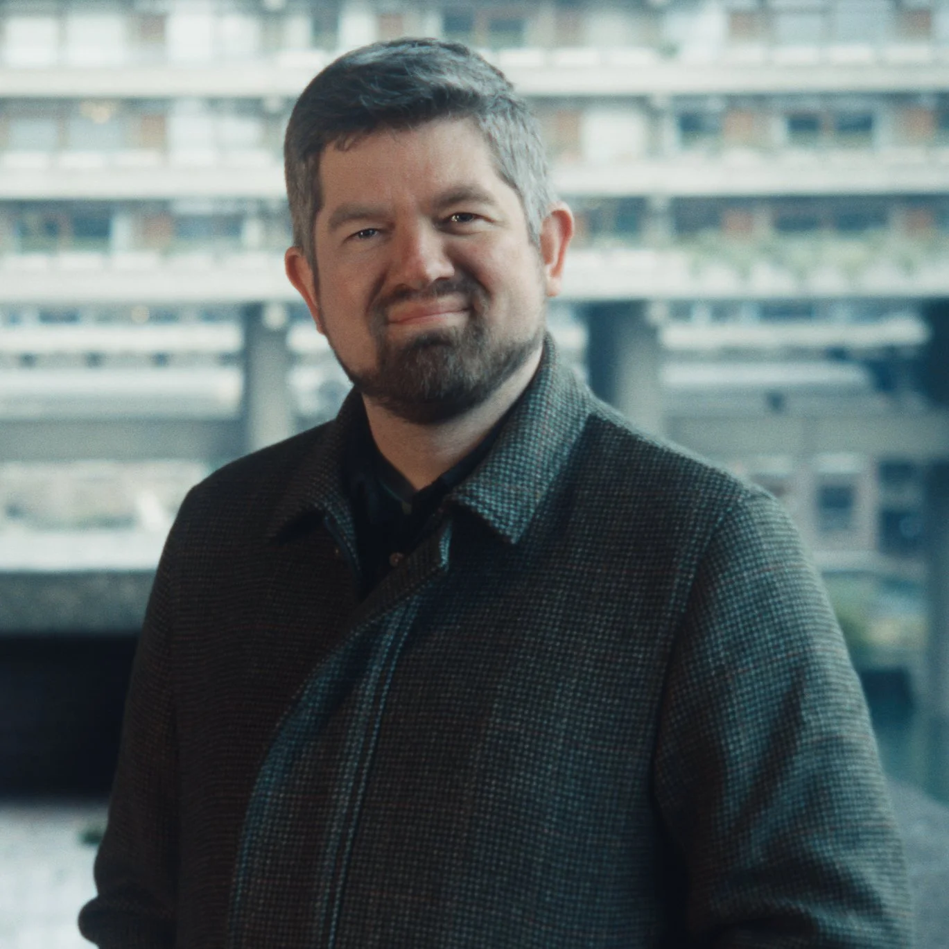 A man with short dark hair and a beard, smiling and wearing a dark checkered blazer, standing indoors with a large window showing a cityscape in the background.