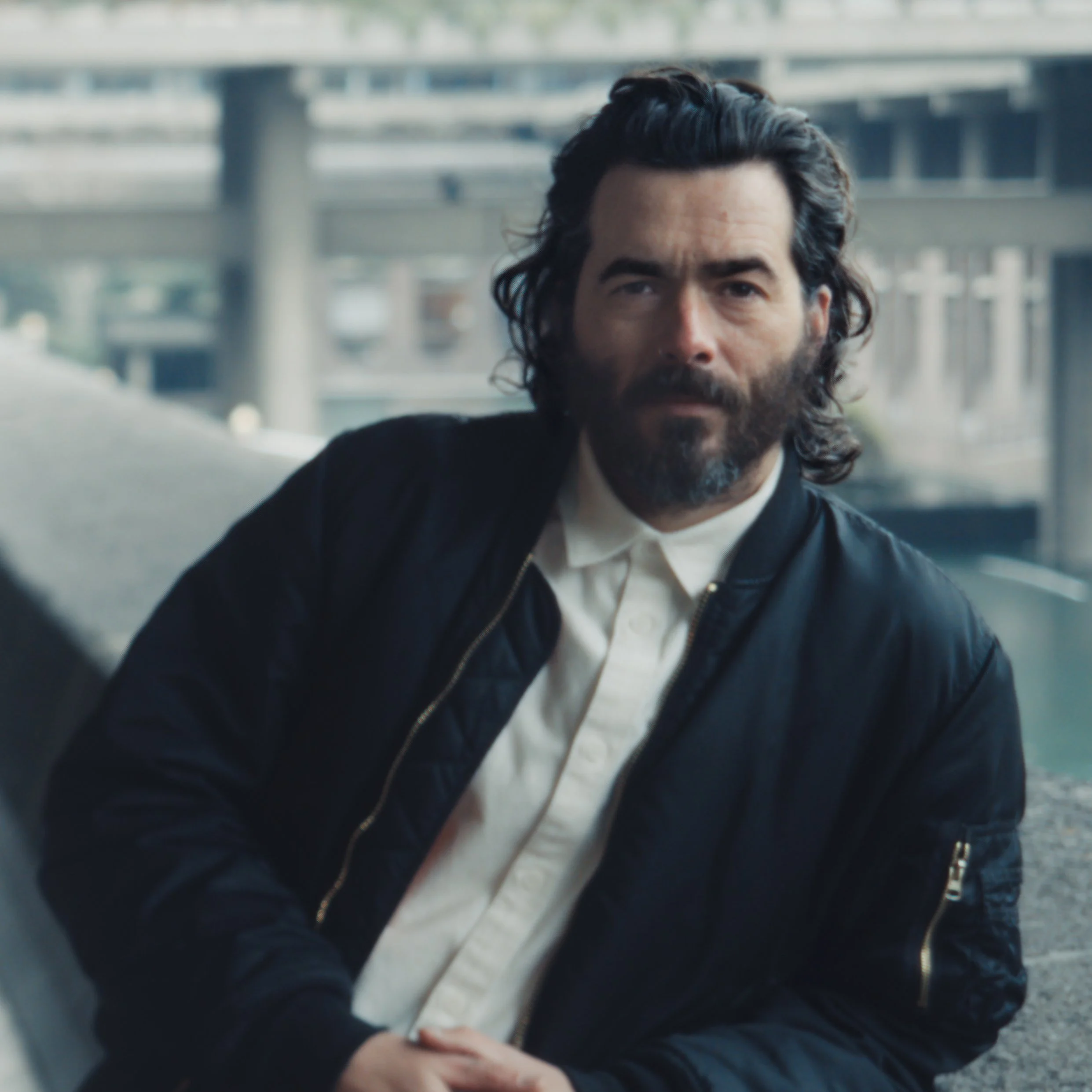 Close-up of a man with dark hair and a beard, wearing a black jacket and white shirt, seen through a reflective surface outdoors.