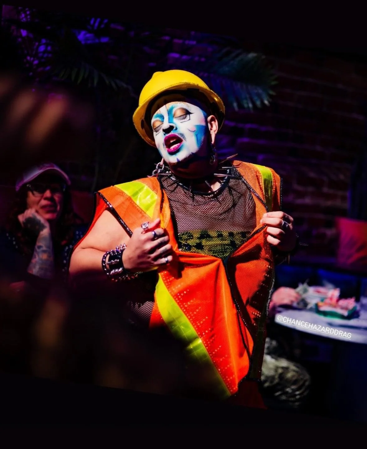 Person with detailed clown face paint wearing a yellow construction helmet, orange safety vest, and black mesh shirt, in a colorful urban environment.