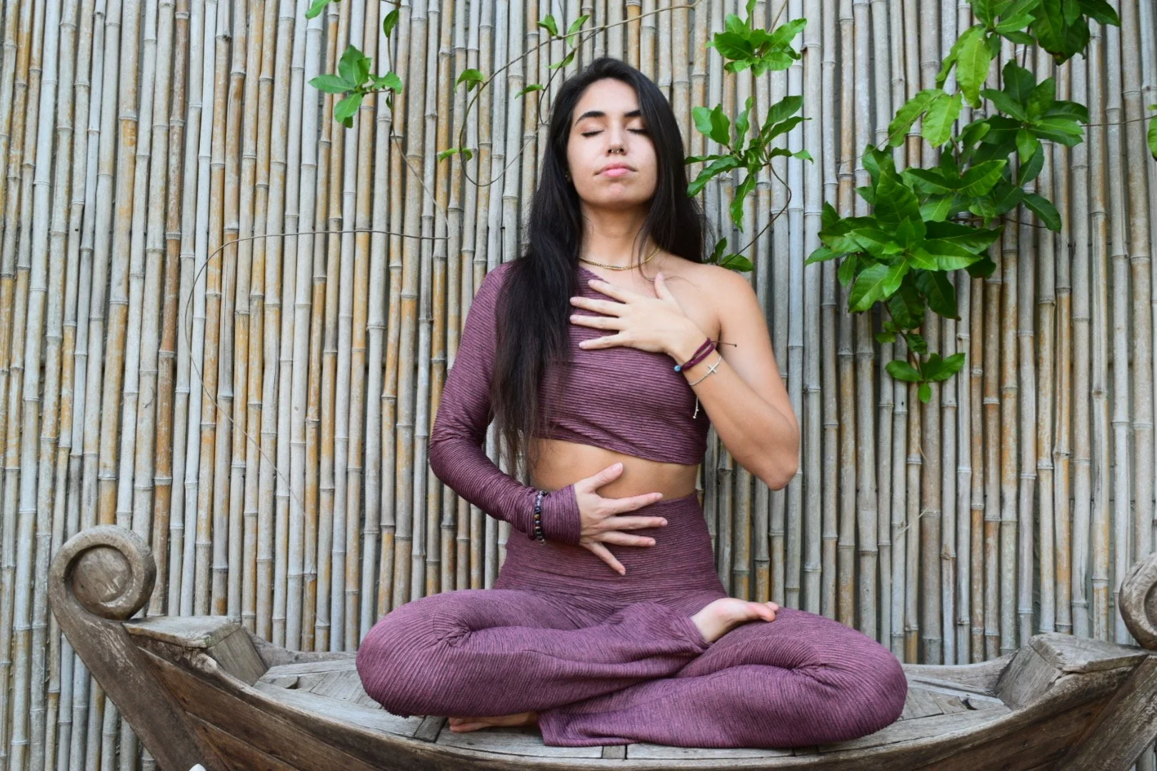 A woman practicing yoga in a meditative pose on a wooden boat-shaped platform, sitting cross-legged with one hand on her chest and the other on her stomach, in front of a bamboo fence with green plants.