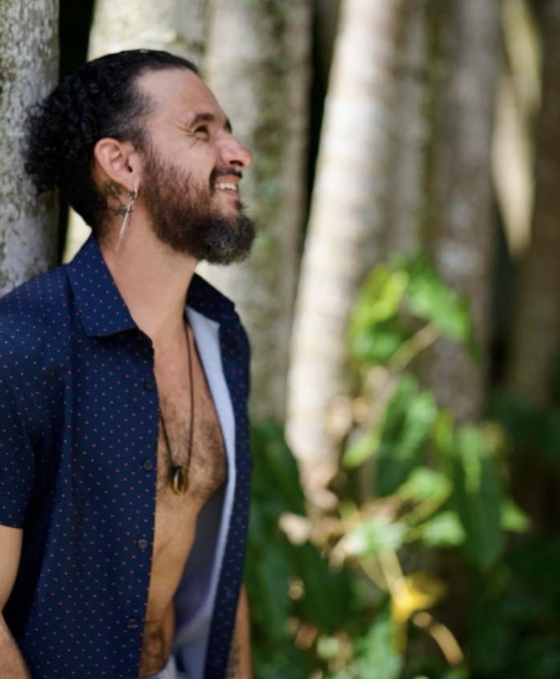 Man with curly hair and beard leaning against a tree, smiling, in a forest setting.