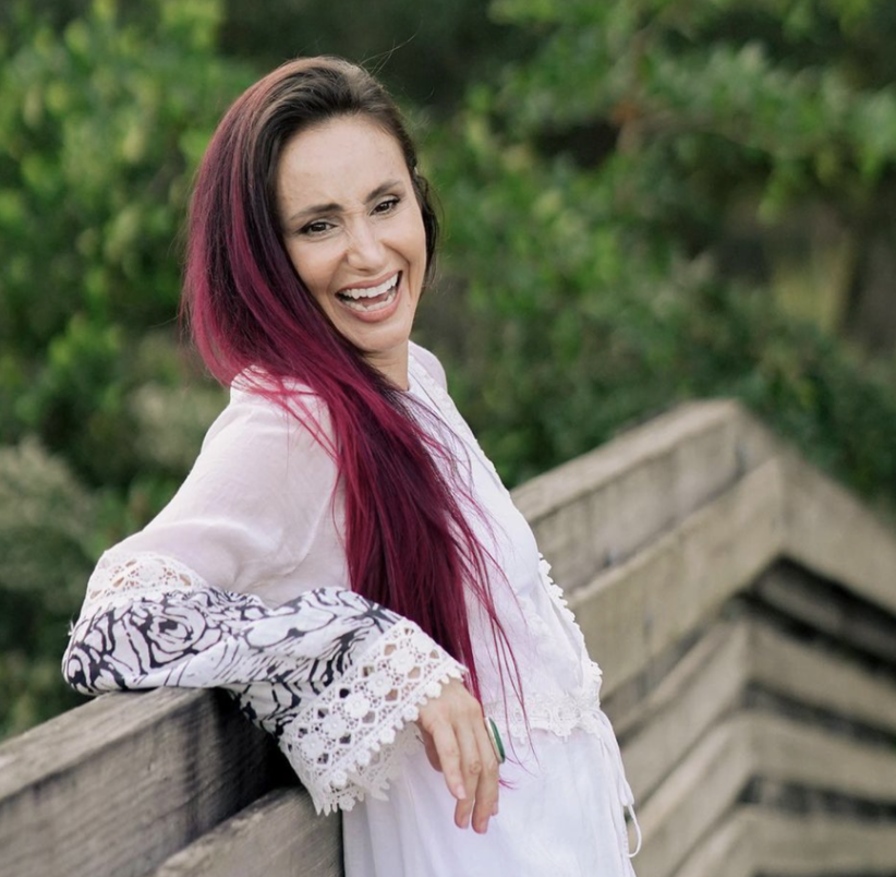 A woman with long brown hair smiling and leaning on a wooden railing outdoors surrounded by greenery.