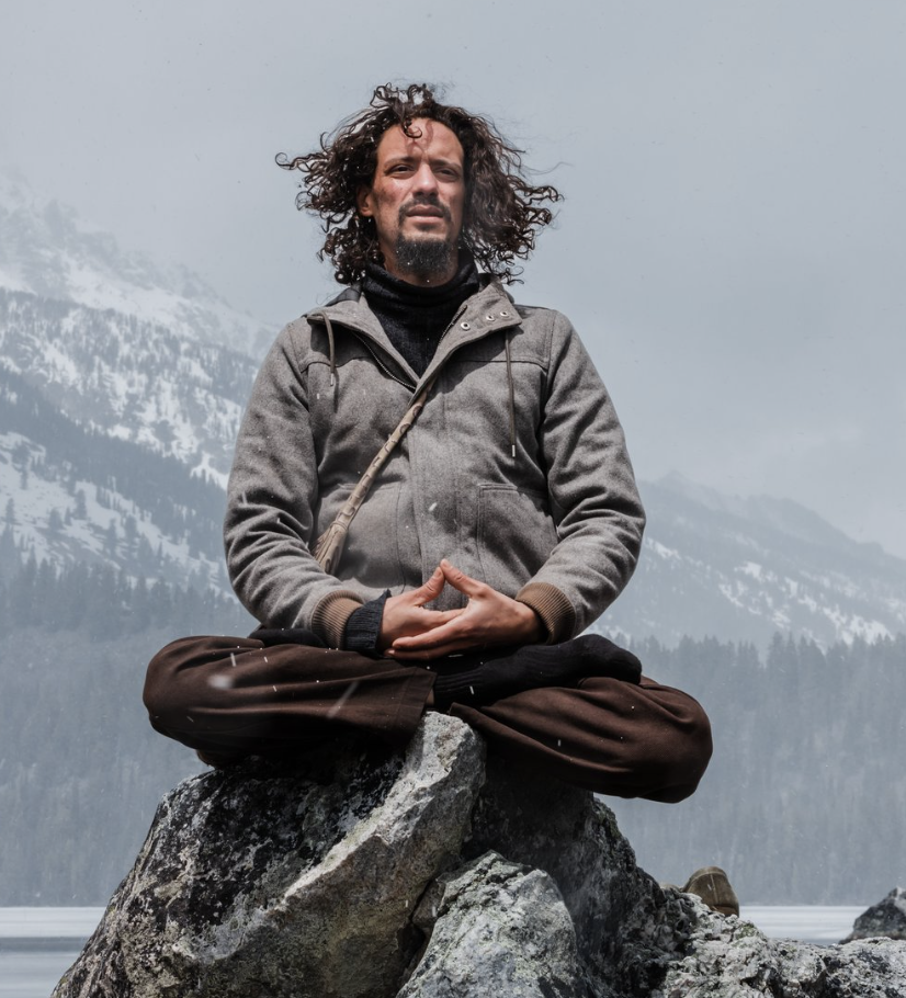 A man with curly hair and a beard meditating on a large rock in a mountainous, snowy landscape during the daytime.