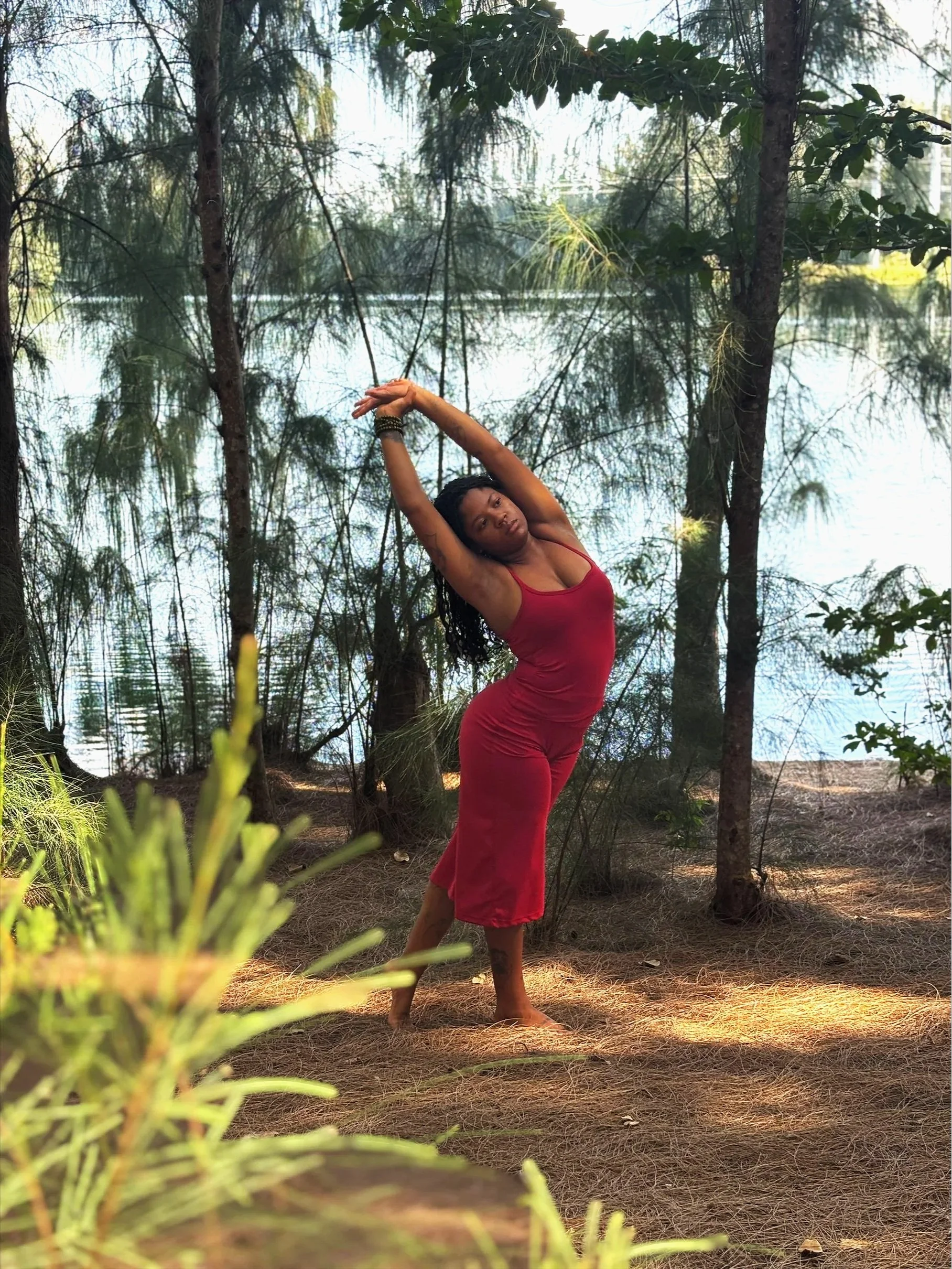 A woman in a red dress practicing yoga outdoors near a lake, standing in a side bend pose with arms raised above her head, surrounded by trees and natural scenery.