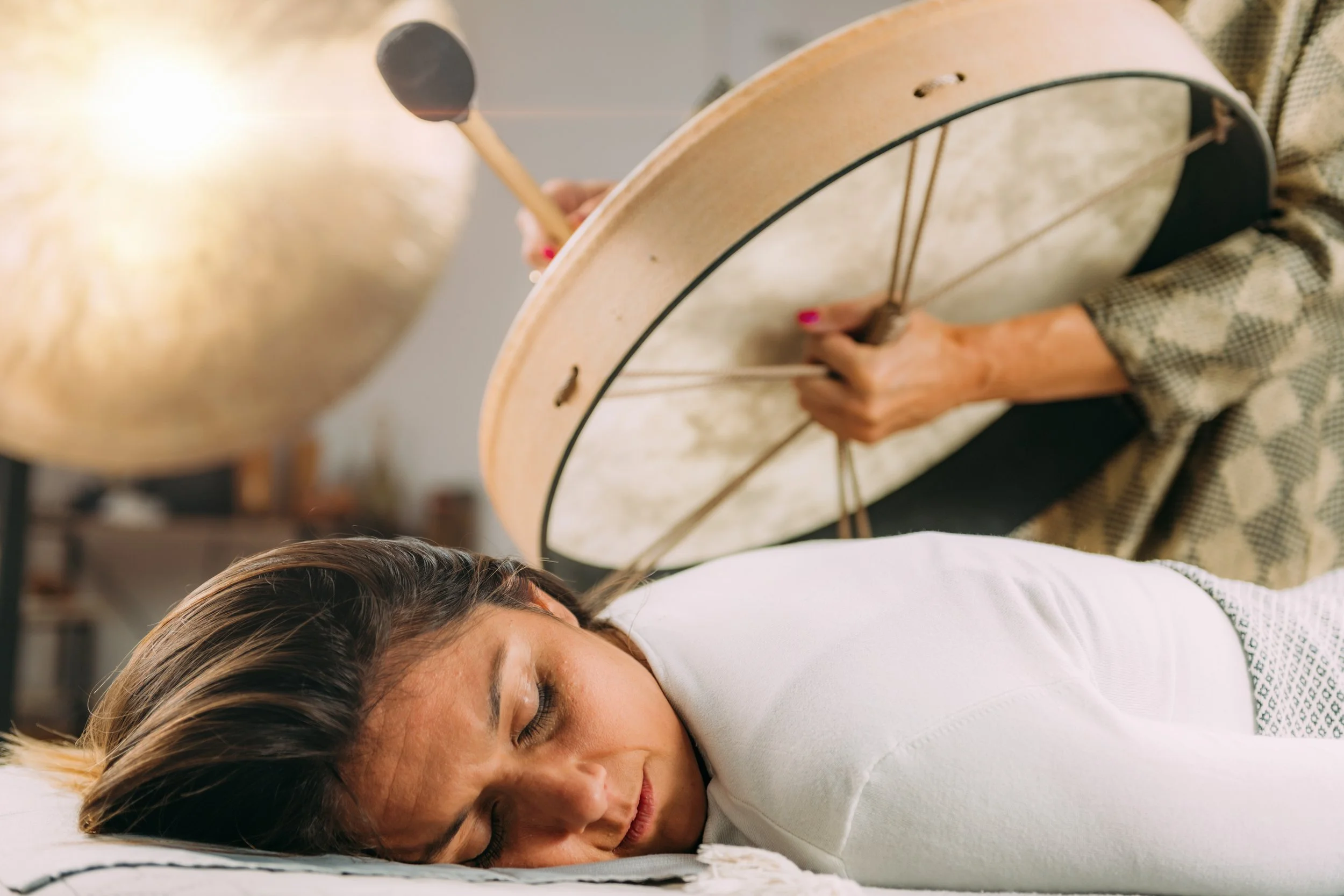 A woman sleeps peacefully on her side with her head on a pillow while a person plays a drum nearby in a cozy room.