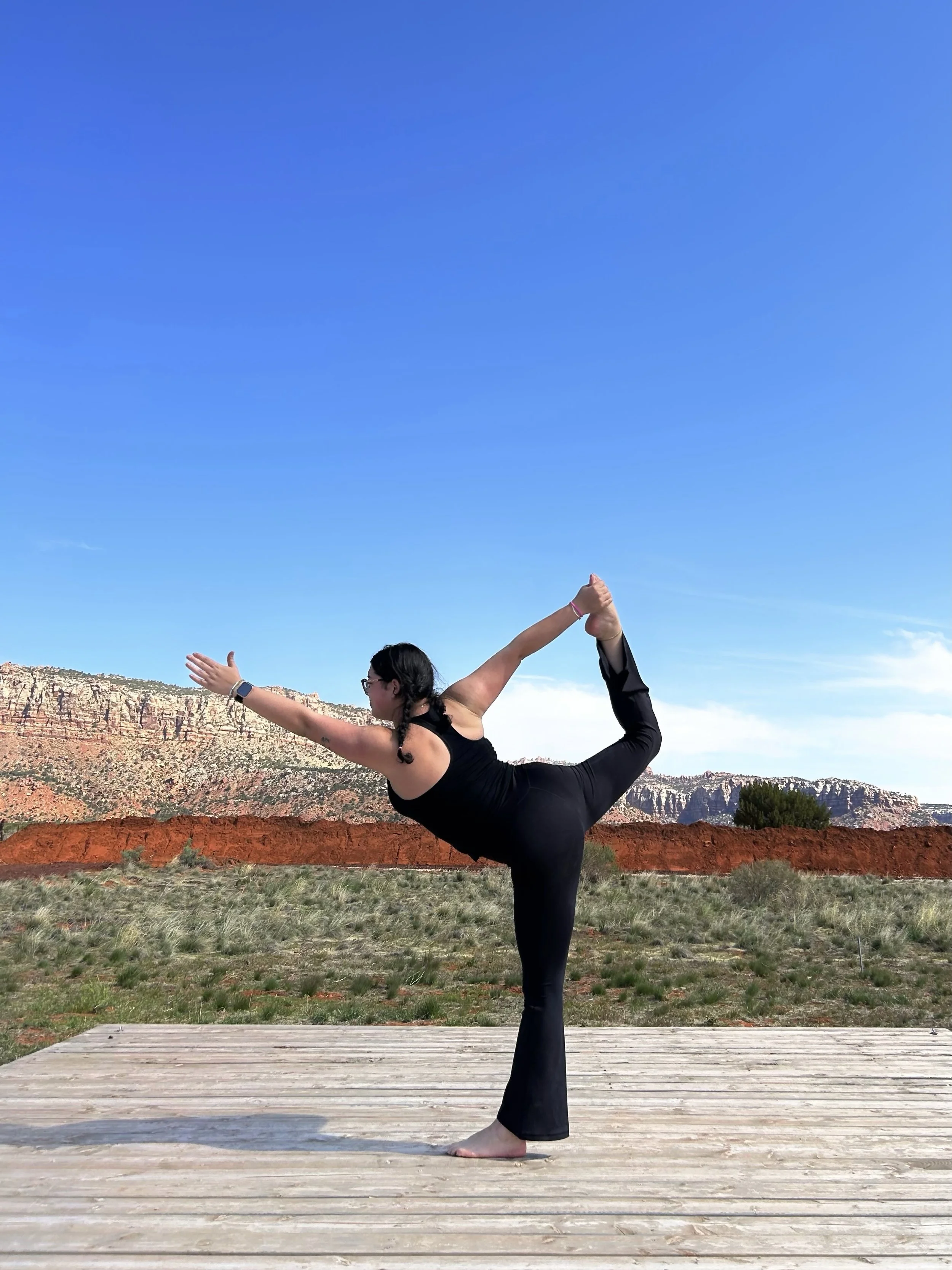 A woman in black yoga attire practicing a balancing pose on a wooden platform outdoors, with red rock cliffs, green vegetation, and a blue sky with some clouds in the background.