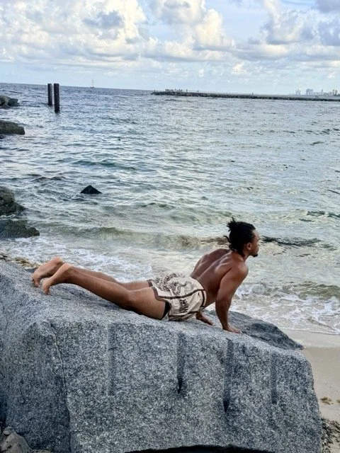 A man with long hair doing yoga on a large rock by the ocean, with a pier and cloudy sky in the background.