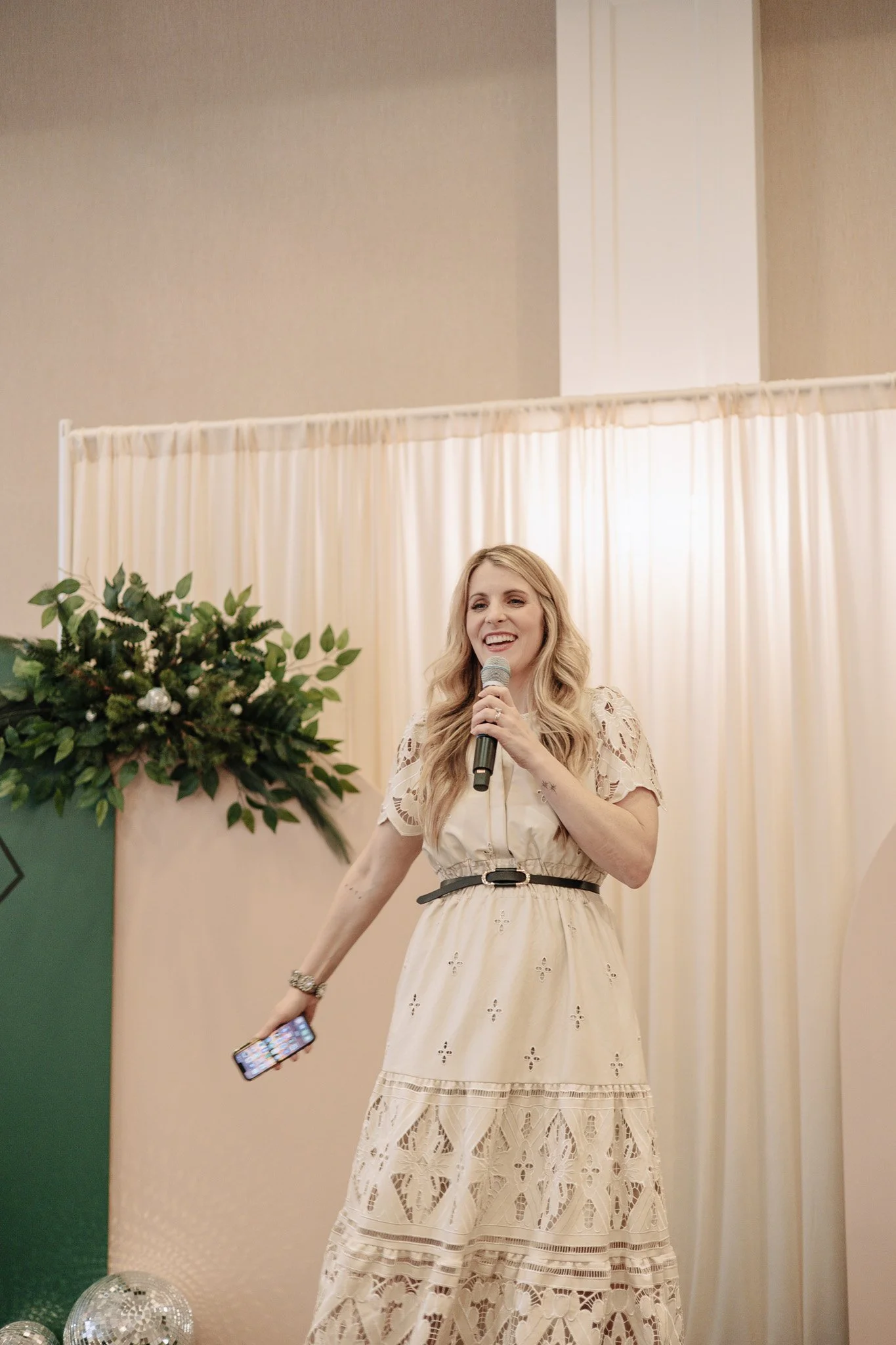 A woman with long blonde hair speaking into a microphone at an indoor event, standing in front of a cream-colored curtain with a large green floral arrangement nearby.