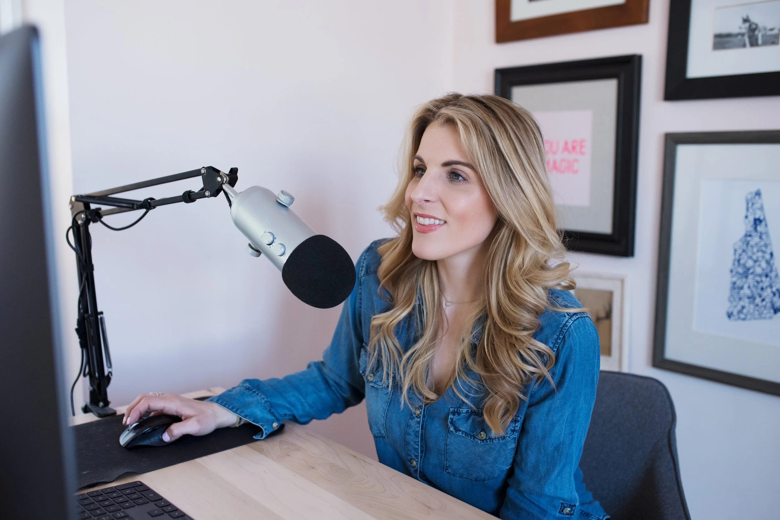 A woman wearing a blue denim shirt speaking into a microphone in front of a computer.