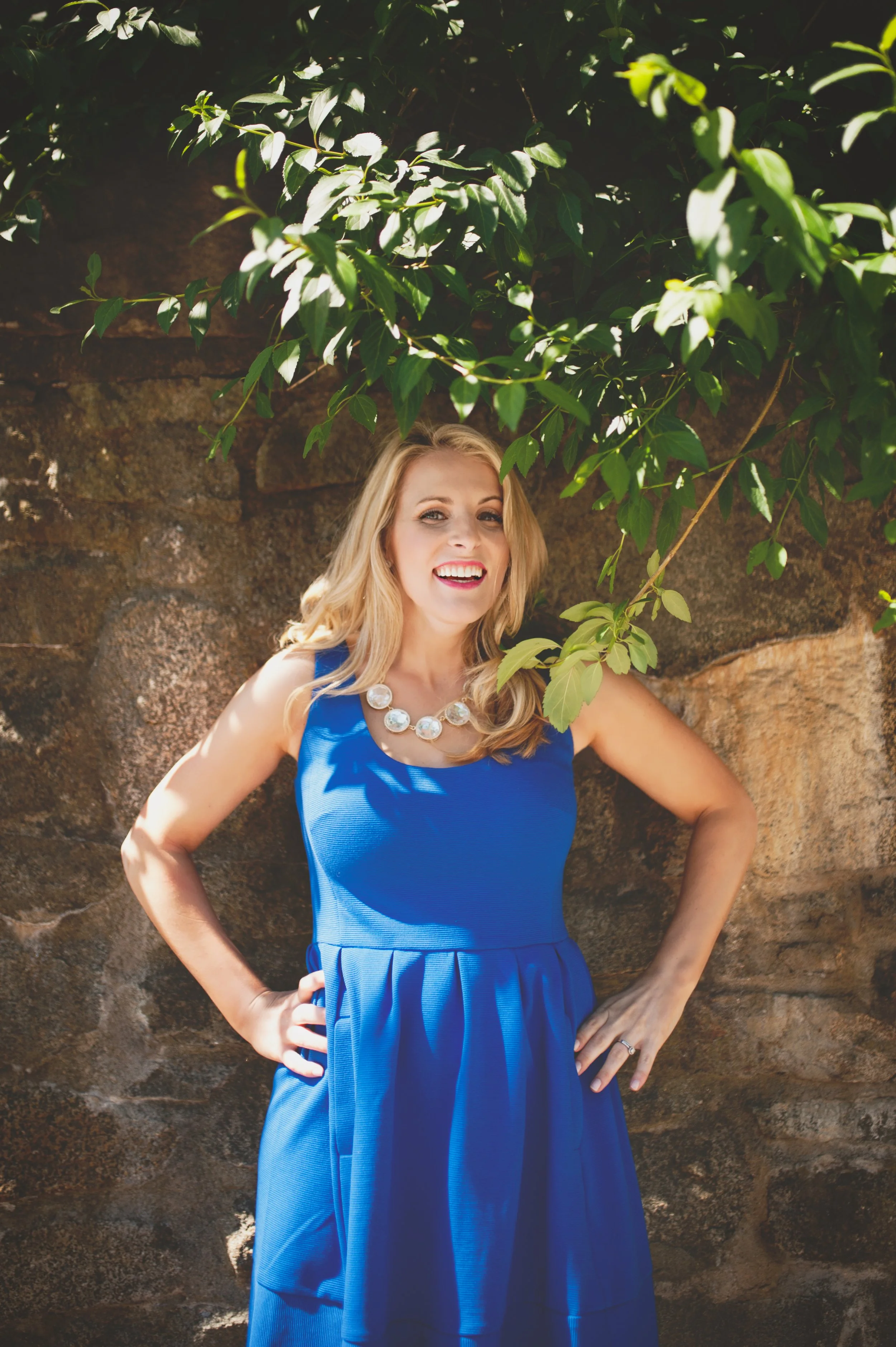 A smiling woman in a blue sleeveless dress with a white necklace, standing outdoors against a stone wall and green foliage.