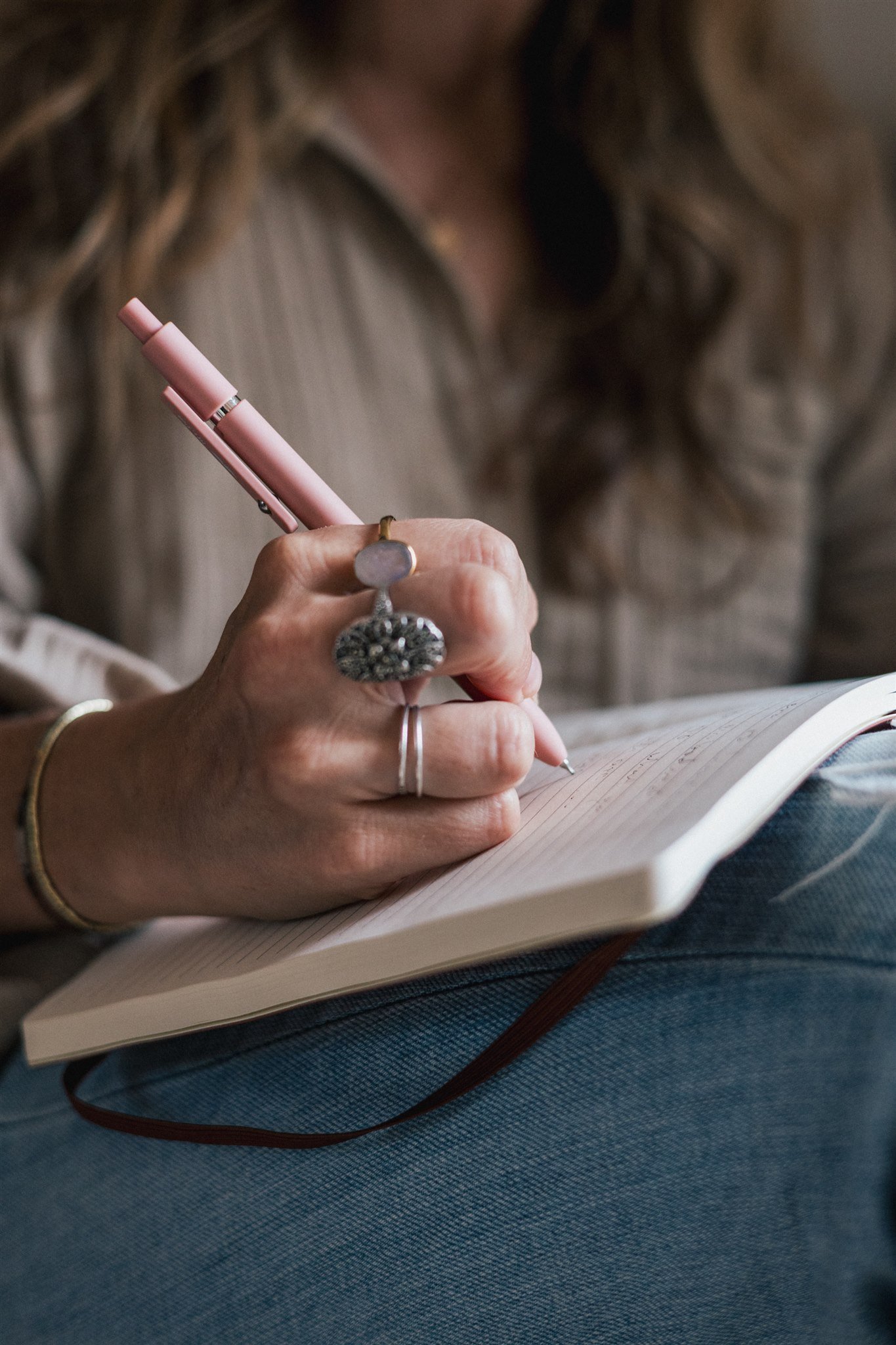 A person writing in a notebook with a pink pen, wearing rings and a bracelet.