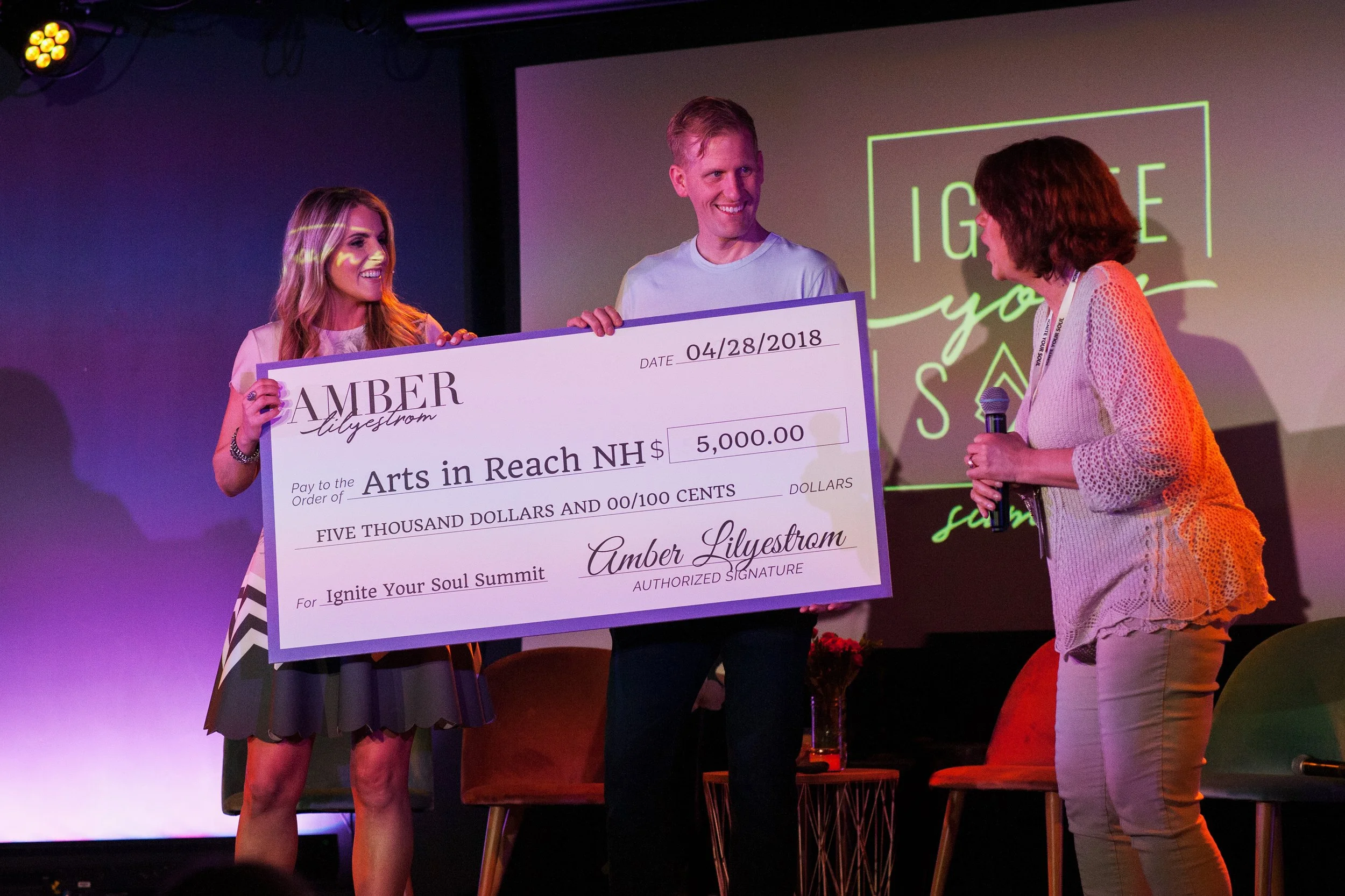Three women on a stage during an award ceremony. One woman is receiving a ceremonial oversized check for $5,000 made out to Arts in Reach NH, from Amber Lyleström, for the Ignite Your Soul Summit. The woman on the right, holding a microphone, is speaking or announcing. In the background, there is a screen with the words 'Ignite your soul' and a floral arrangement on a small table.