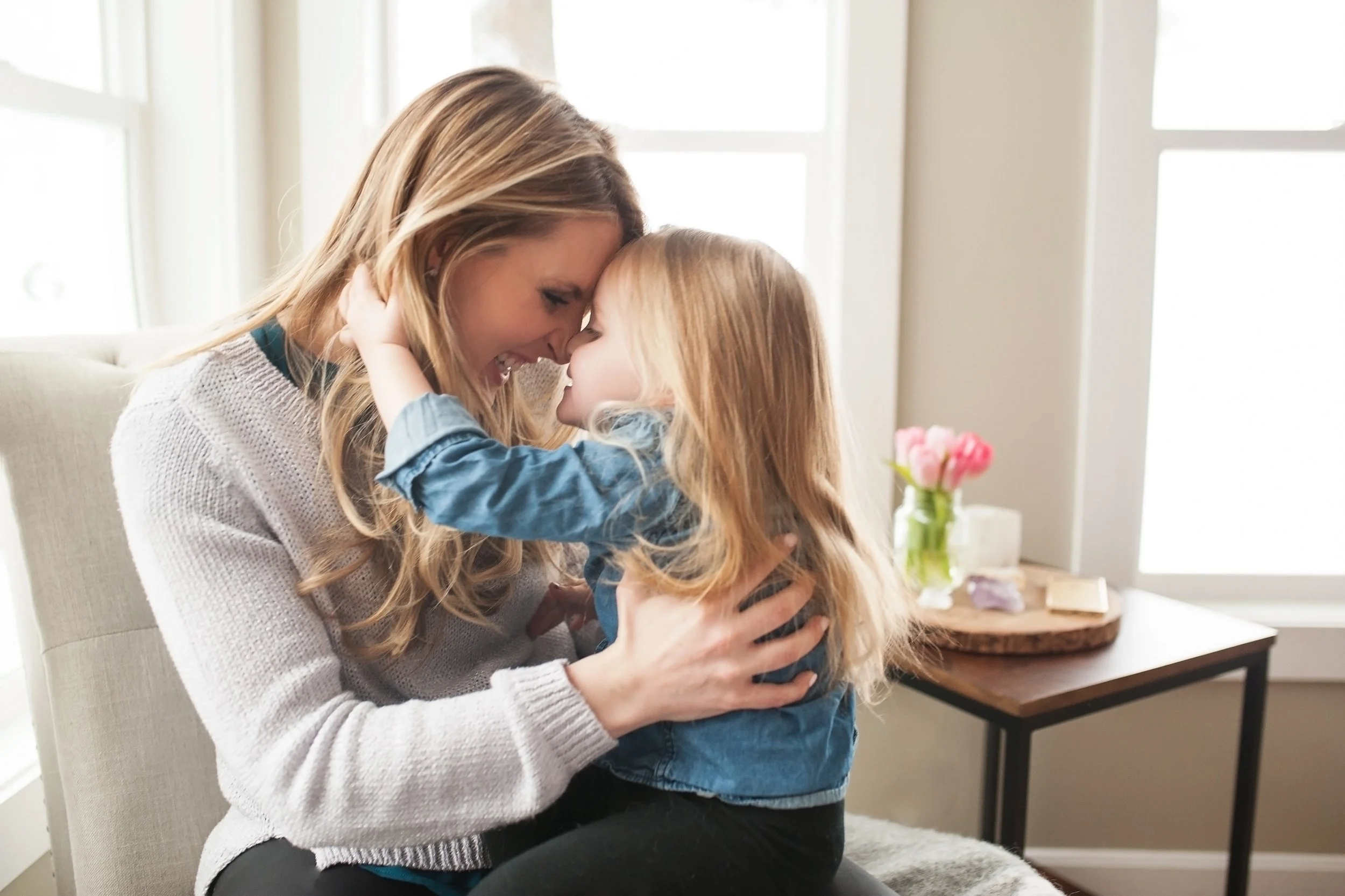 A woman and a young girl are touching foreheads and smiling affectionately indoors with a window and a table with pink flowers in the background.