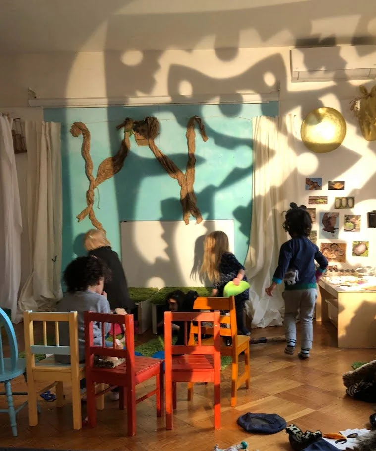 Children playing indoors with shadows of tree branches cast on the wall, colorful chairs, and artwork displayed on the right wall.