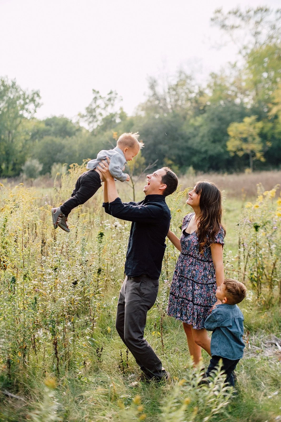 A happy family outdoors in a field; a man lifts a young child into the air while a woman and another young boy look on smiling.