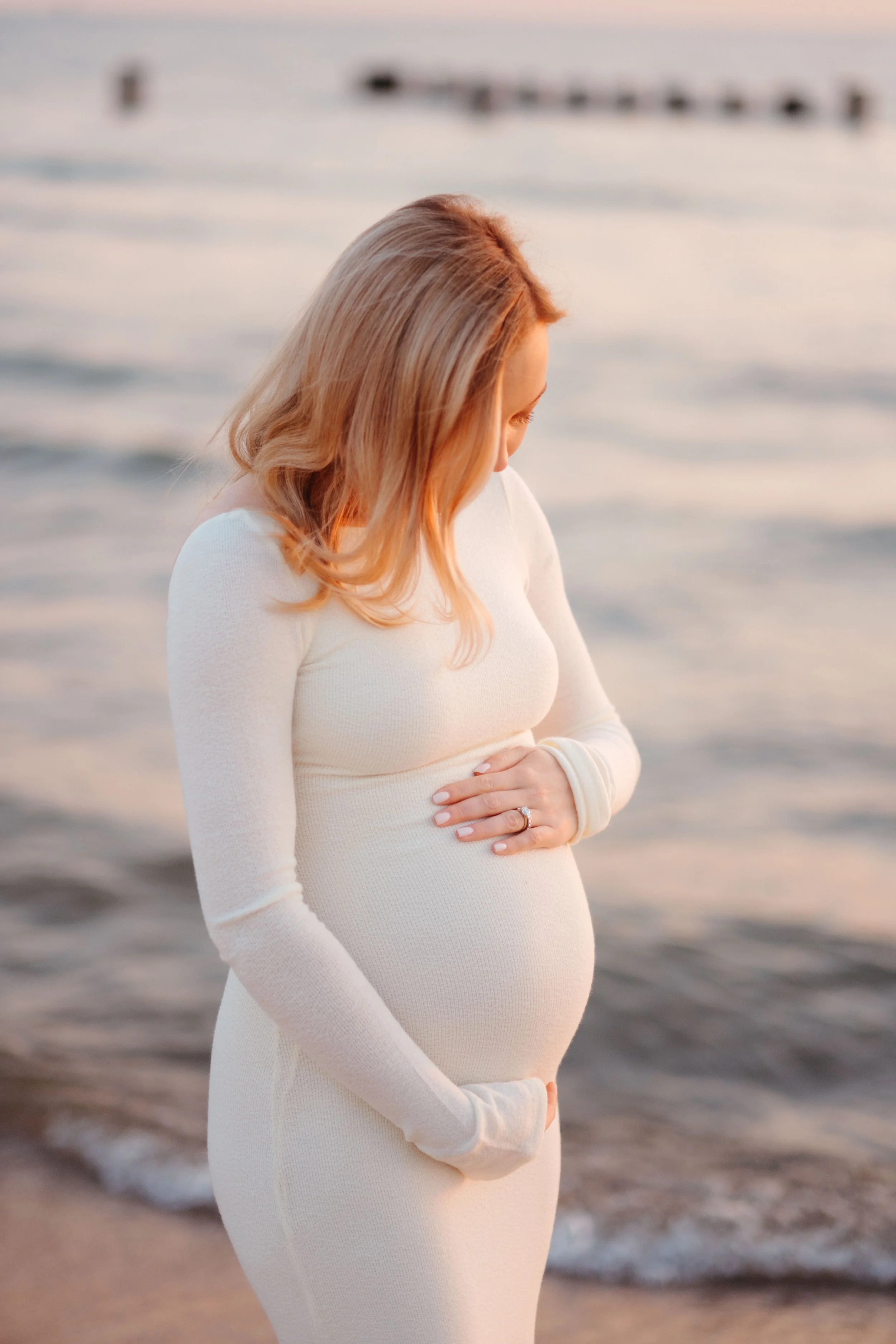 A pregnant woman in a white dress standing on a beach at sunset, looking down at her belly with her hand resting on her stomach.