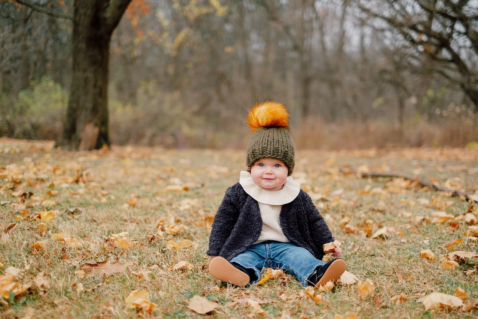 A baby girl sitting on the ground in a park surrounded by fallen autumn leaves, wearing a knit hat with a pom-pom, a sweater, and a jacket, during daytime.