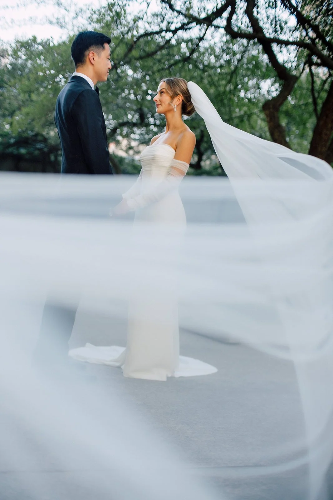 A bride and groom standing face to face outdoors with trees in the background. The bride is wearing a white wedding dress and veil, the groom is in a black suit, and they are holding hands.