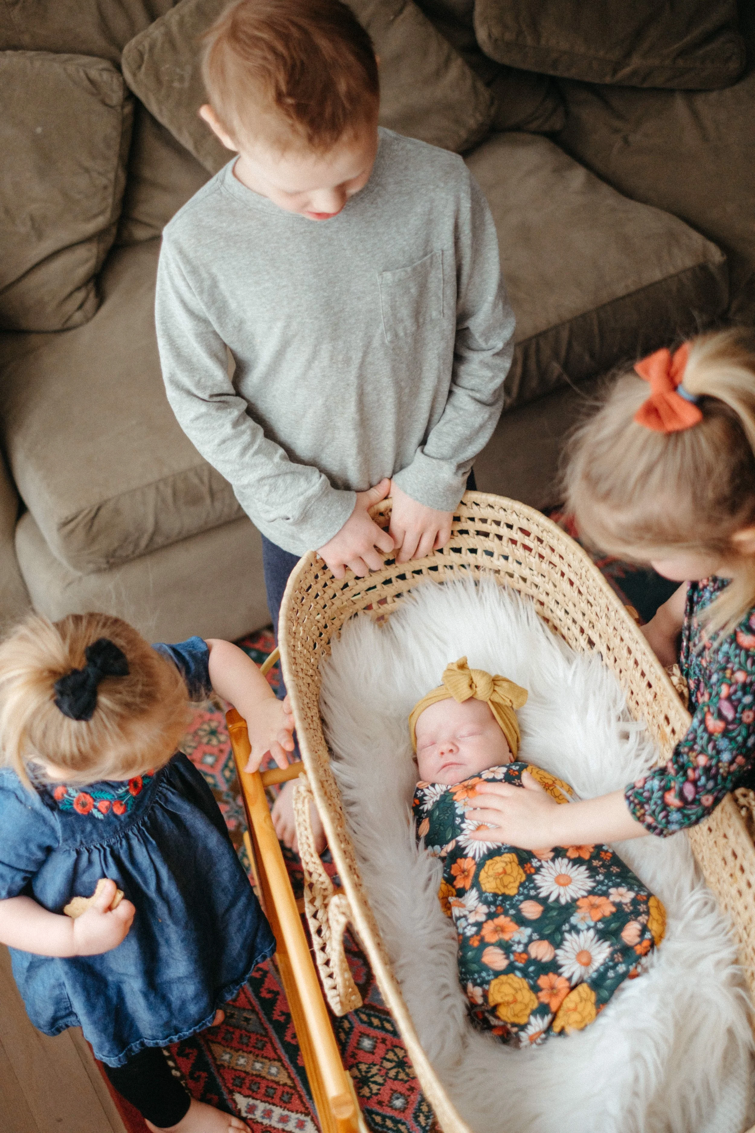 A young boy and two young girls look at a sleeping baby girl in a wicker bassinet. The baby wears a floral dress and a yellow headband, while the girls wear colorful dresses and the boy wears a gray long-sleeve shirt. They are indoors on a patterned rug.
