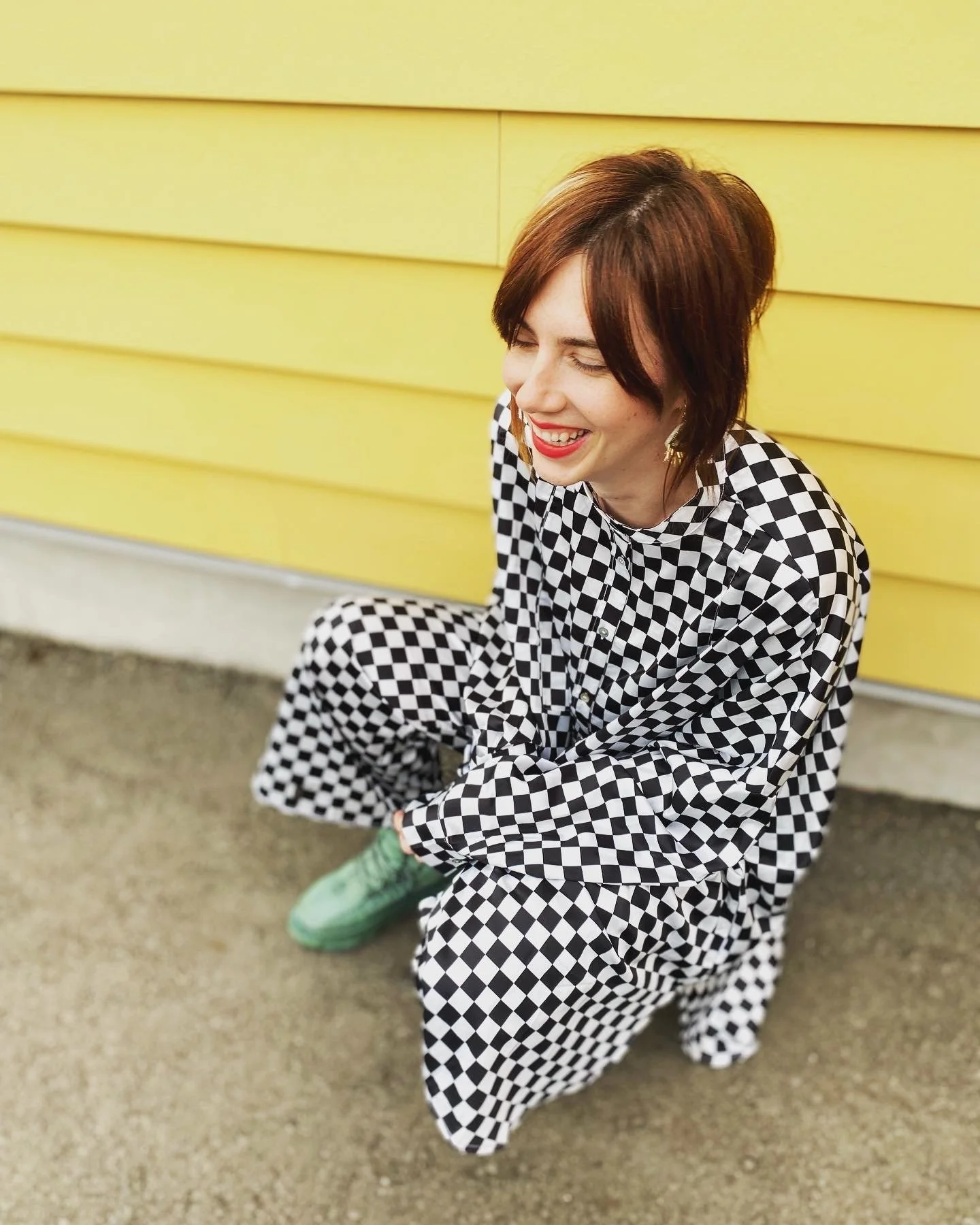 A woman with short brown hair is laughing and crouching against a yellow wall, wearing a black and white checkered outfit and green shoes.