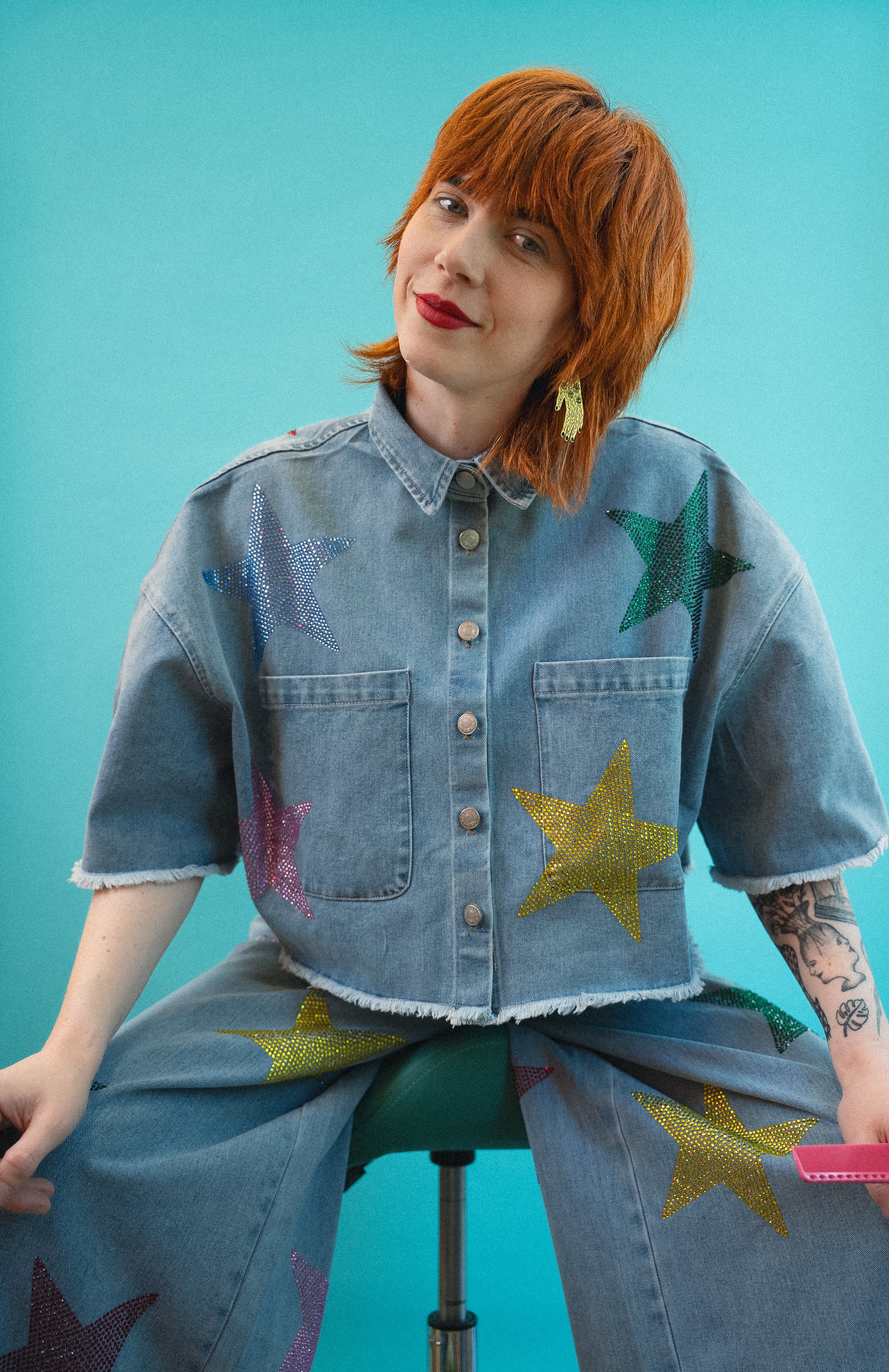 Rebecca Raine is the owner of Lady Neptune and she is a master at giving haircuts seattle can't stop talking about. Known for her expressive fashion choices, this picture shows her with a teal backdrop wearing a denim outfit with stars.