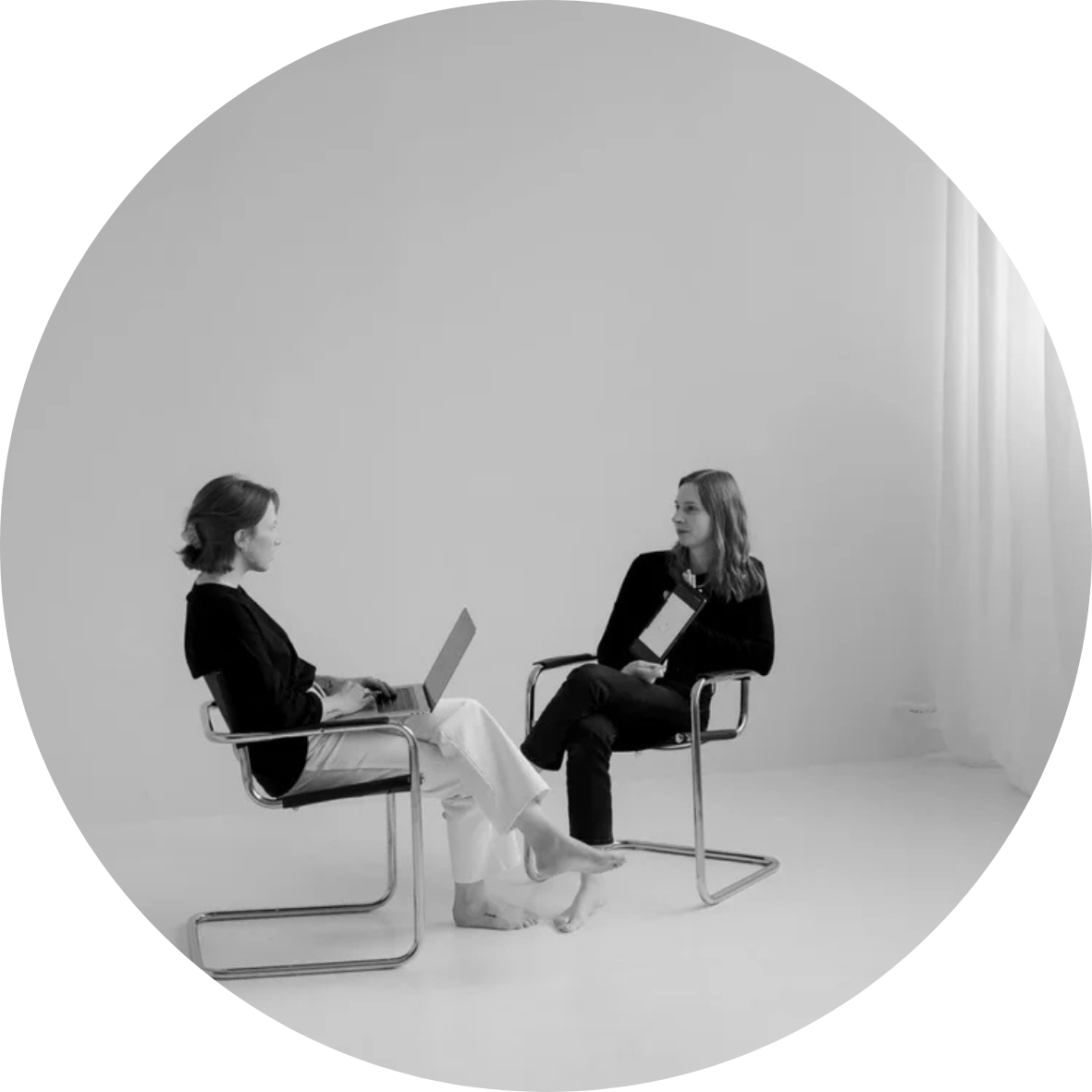 Two women sitting across from each other in a minimalist studio, engaged in conversation, one holding a tablet and the other using a laptop.