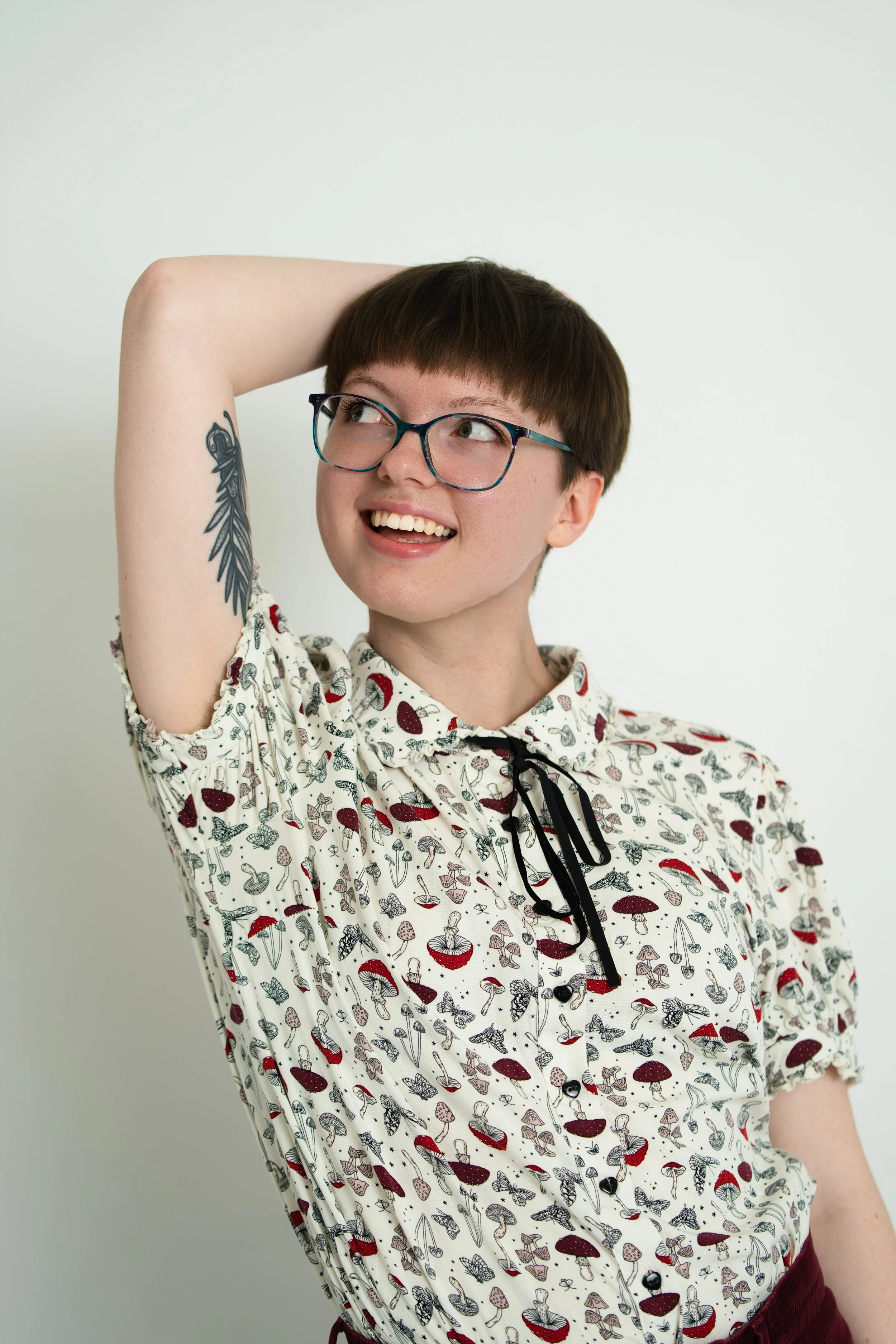Elizabeth has short brown hair and blunt bangs above their eyebrows. They are wearing a mushroom patterned button up, multicolor eyeglasses, and a spoon tattoo is visible on their upper arm.